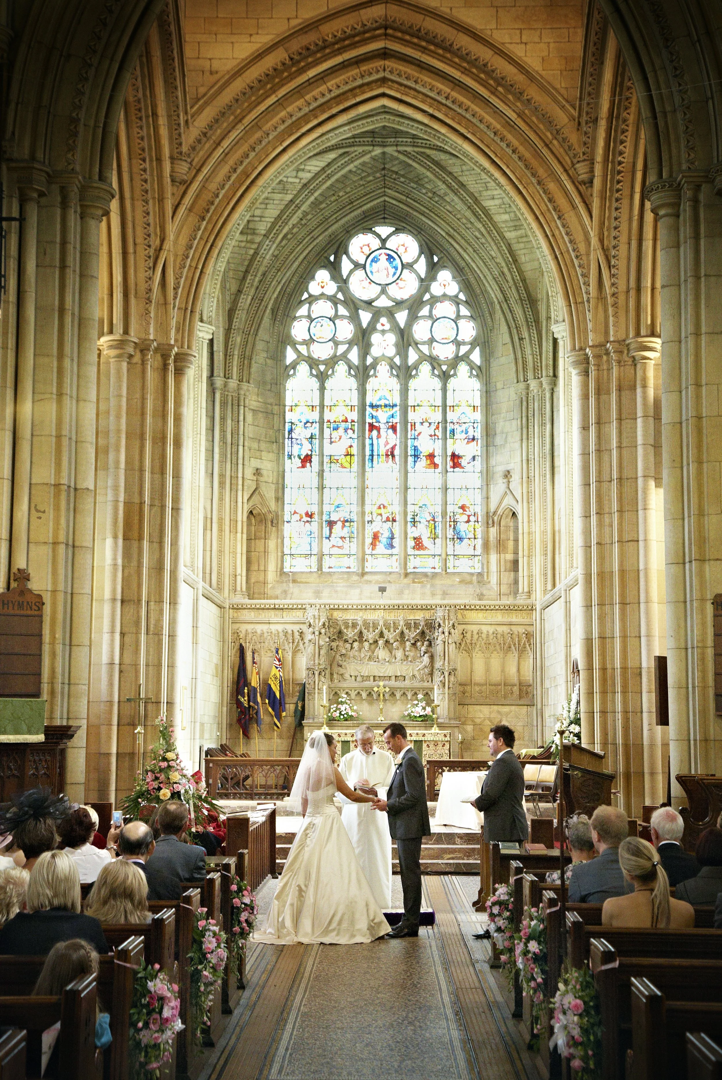 A wedding ceremony inside a church with stained glass windows and gothic architecture. The bride and groom are standing before a priest, exchanging vows, while the officiant looks on. Guests are seated in pews decorated with pink flowers, and the cou