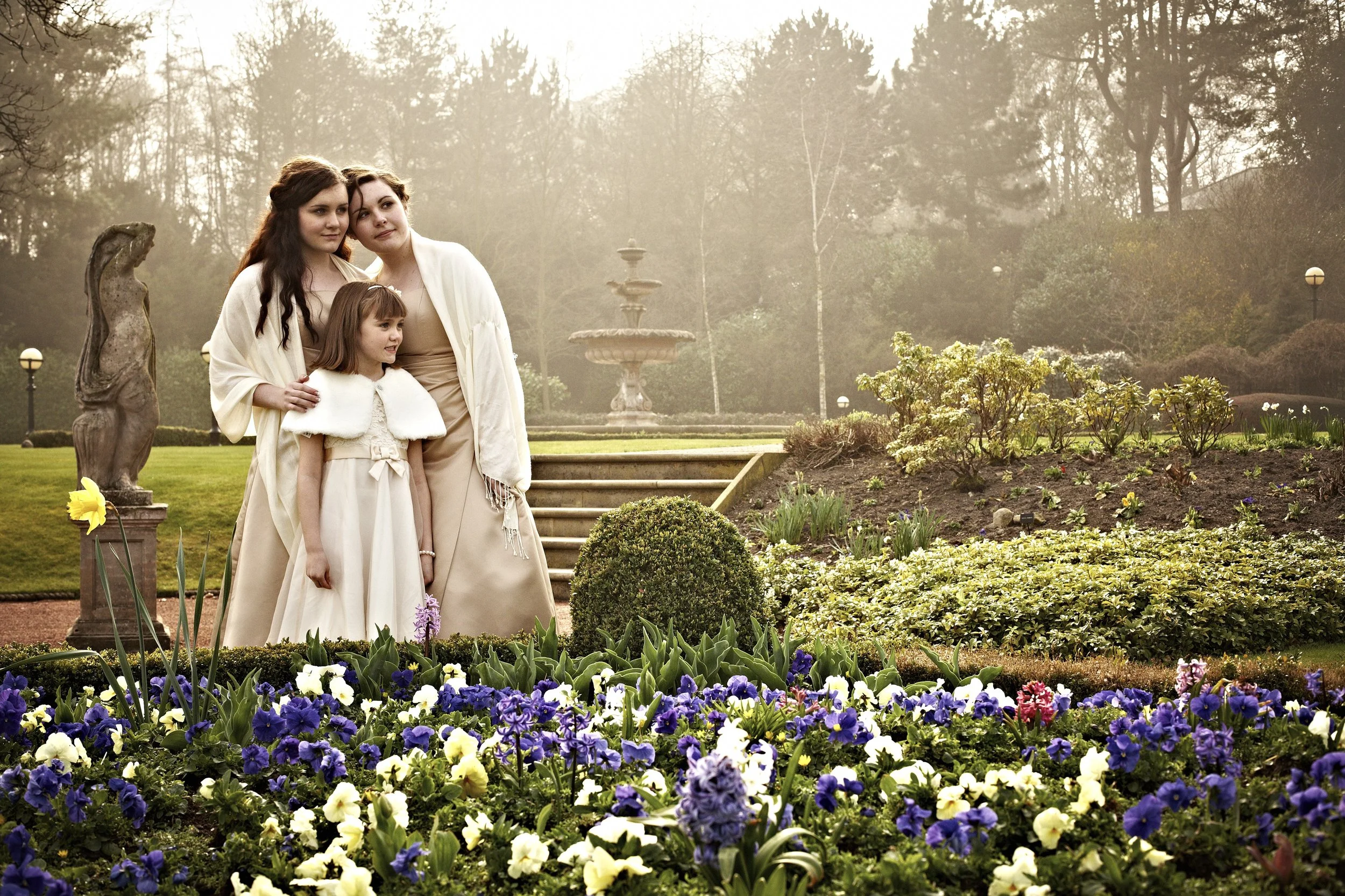 A woman with two young girls standing together in a garden with colorful flowers in the foreground, a fountain in the background, and trees and lamp posts around.
