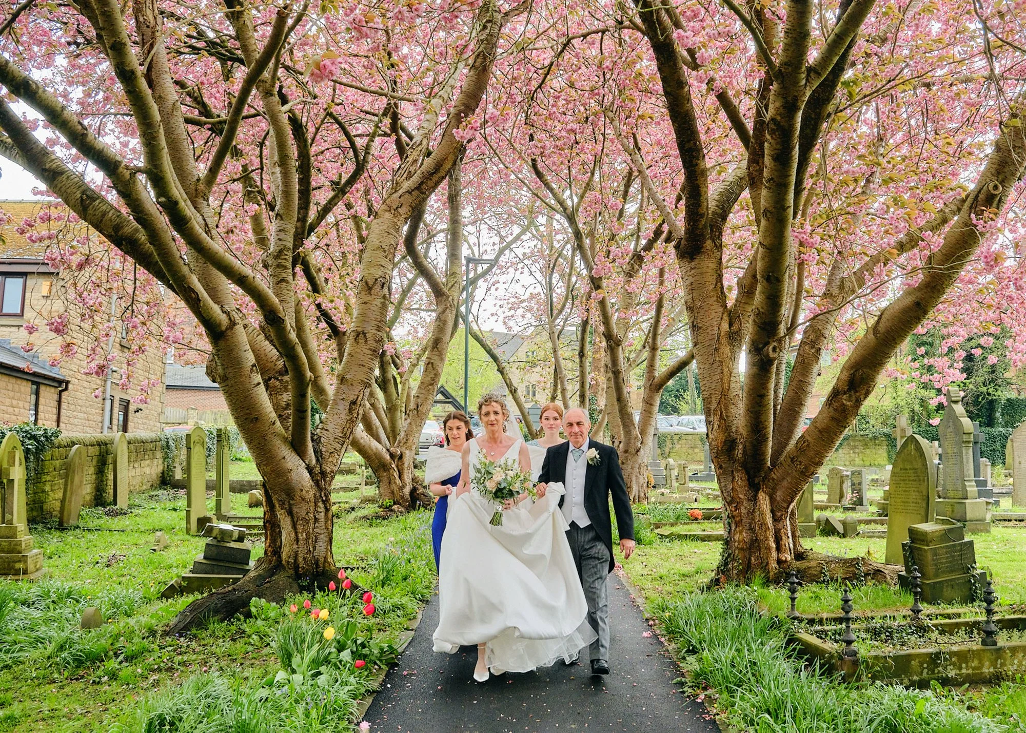 A bride and groom walking hand in hand through a cemetery with flowering trees and historical headstones during a wedding ceremony.