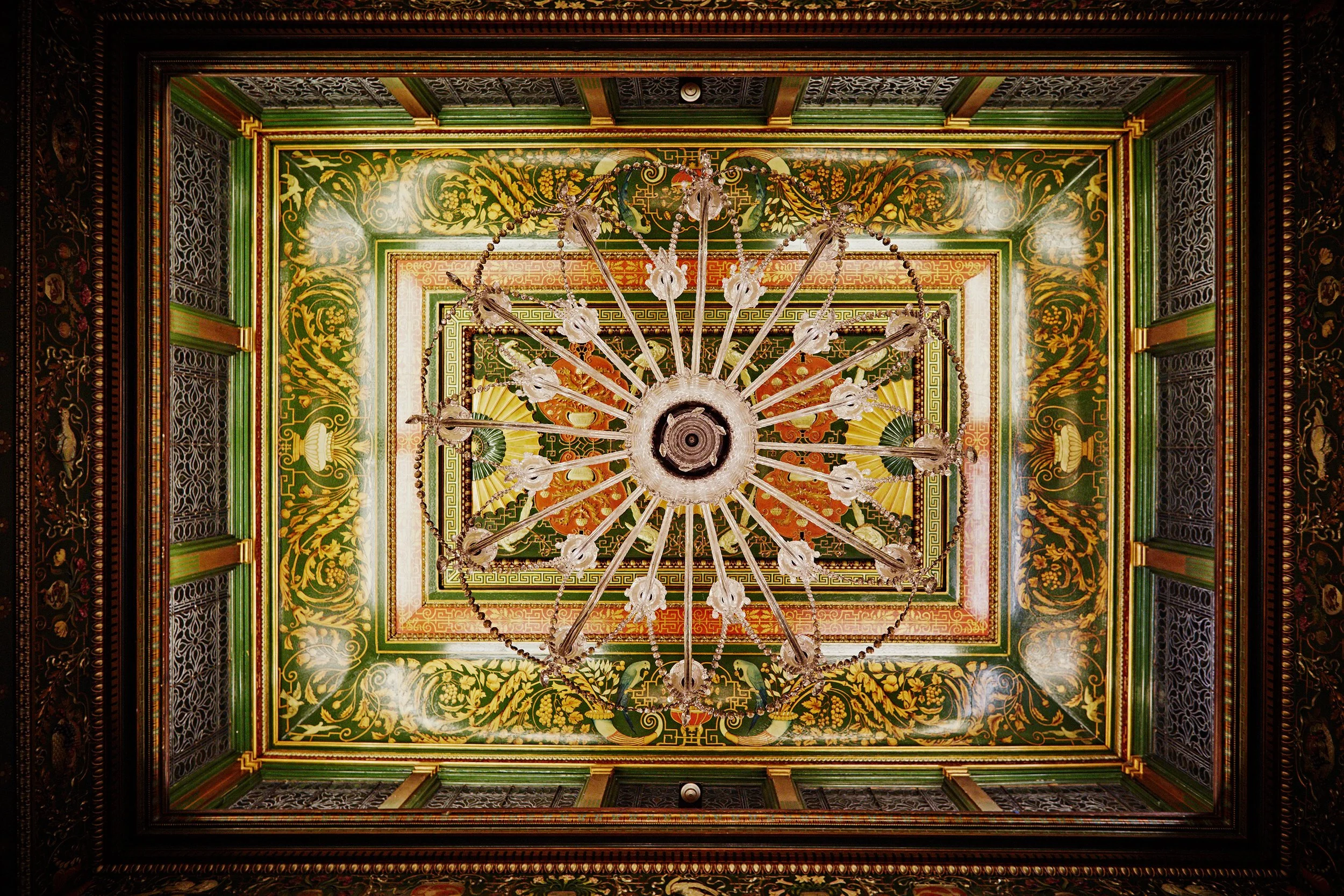 Looking upward at a large ornate chandelier hanging from a decorated ceiling with intricate gold, green, red, and black patterns, surrounded by detailed framing and woodwork.