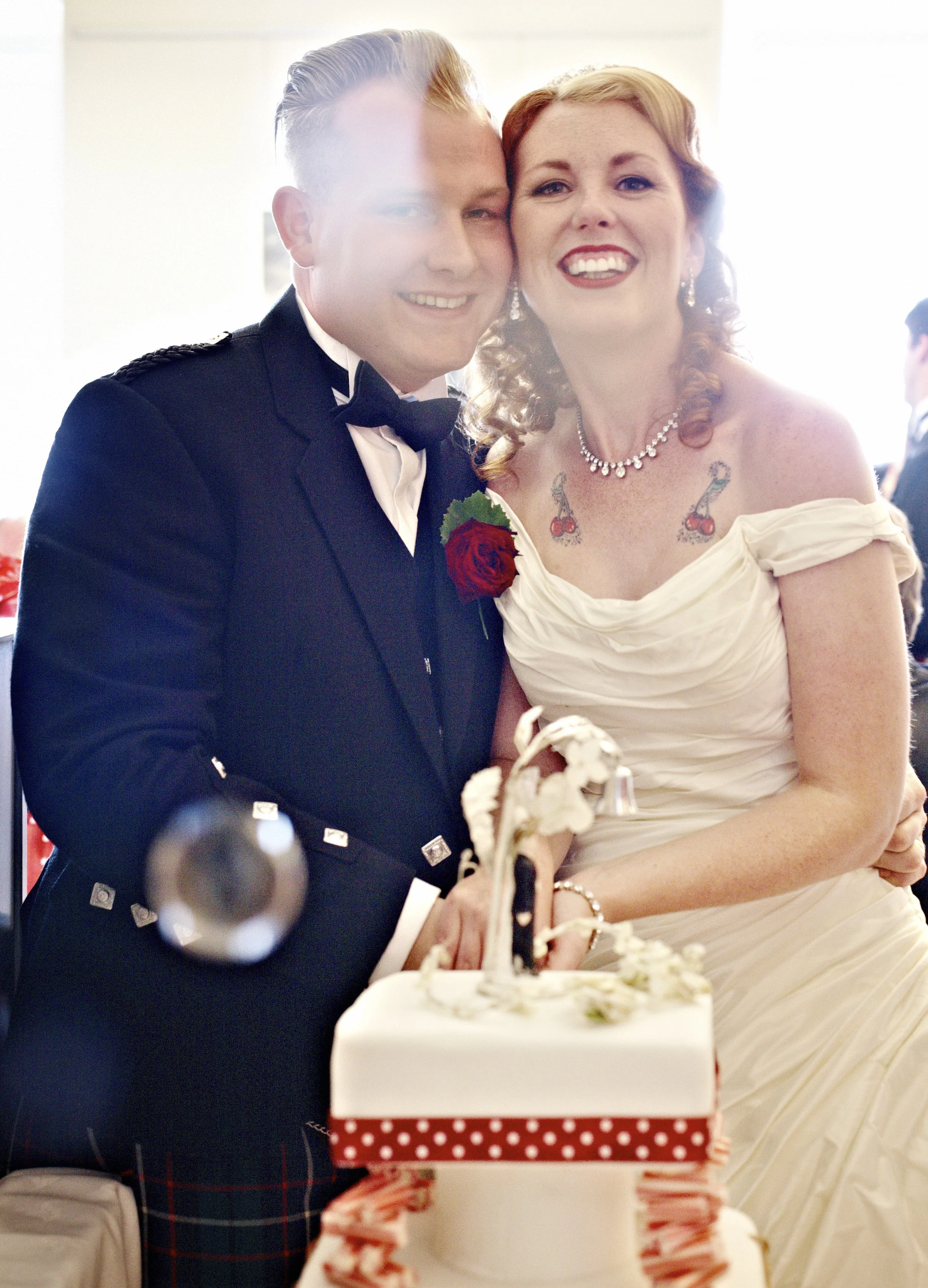 A smiling couple at a wedding with a cake in front of them, the man dressed in a tuxedo with a red rose boutonniere, the woman in a wedding gown with tattoos and jewelry, celebrating.