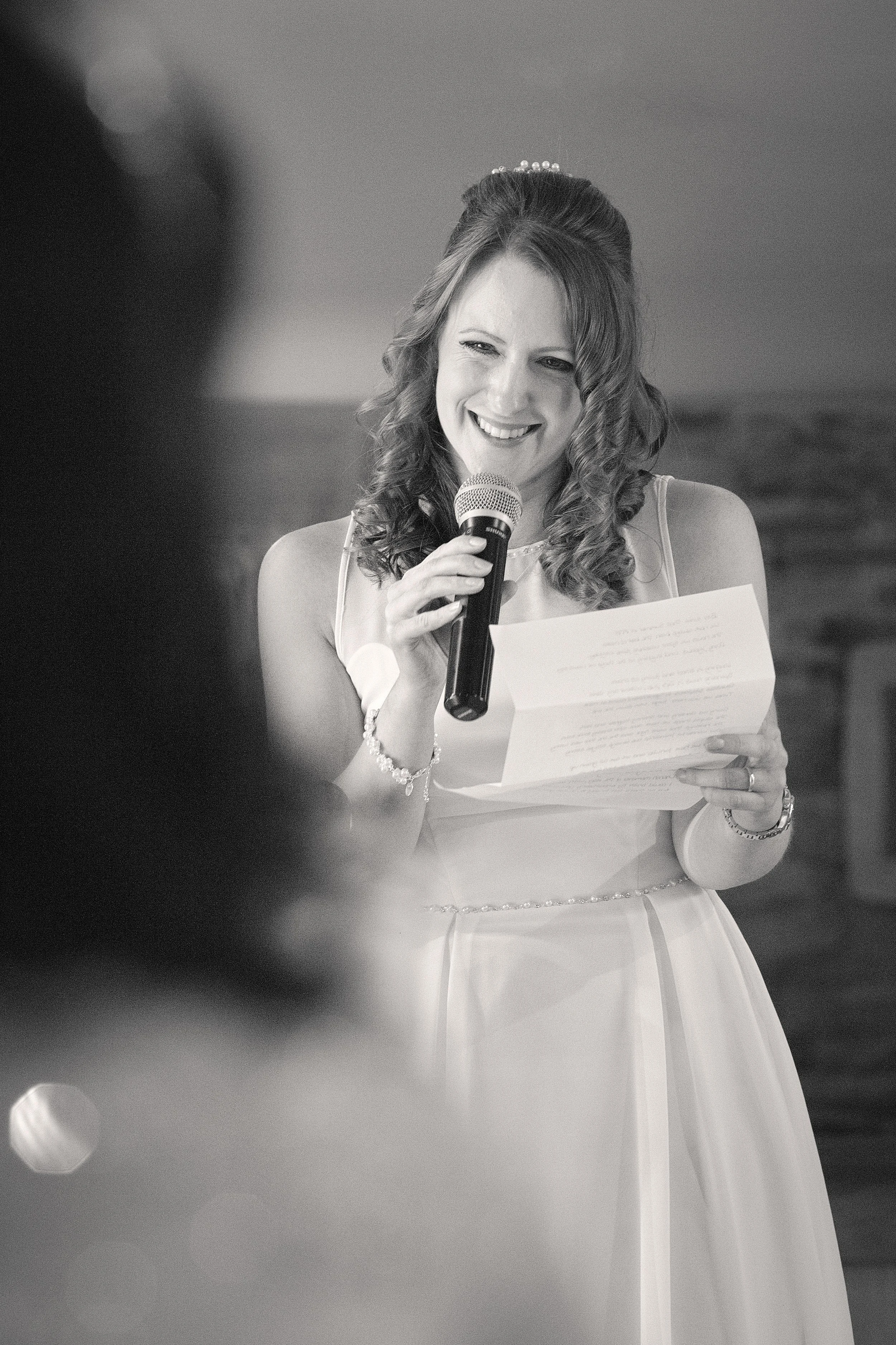 Black and white photo of a smiling woman in a white dress holding a microphone and reading from a paper, likely at a formal event or wedding.