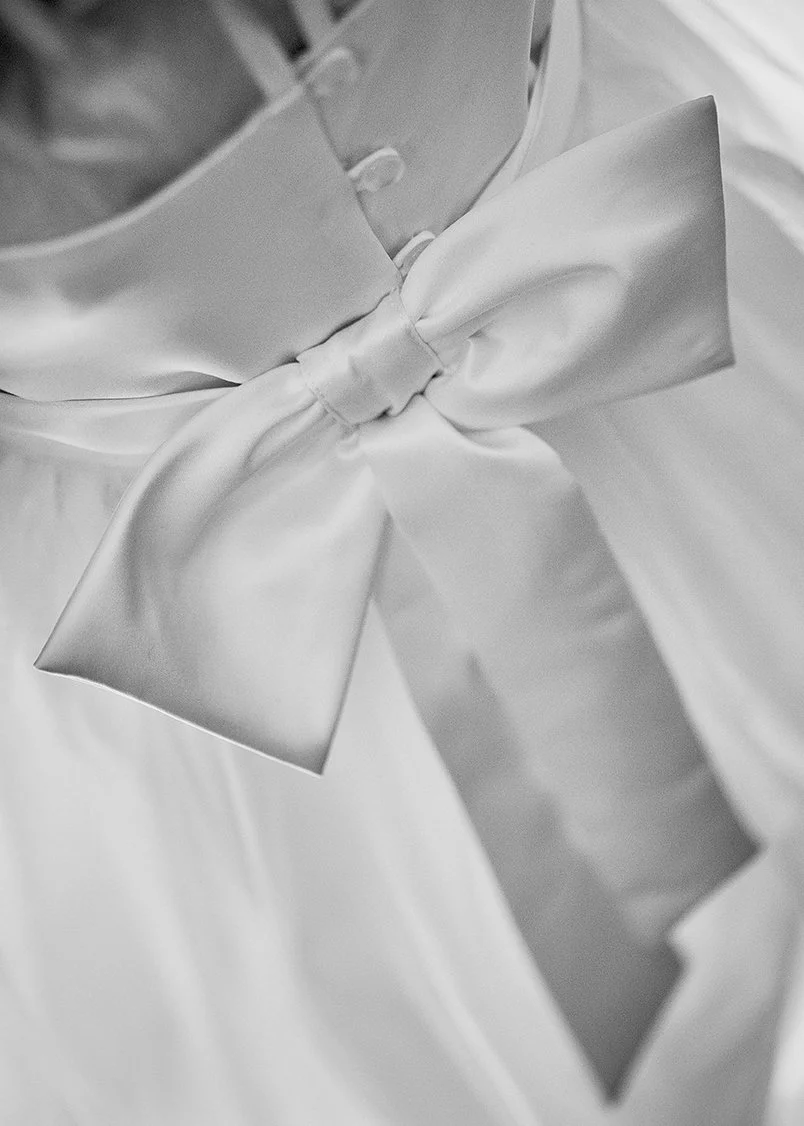 Close-up of a satin bow tied on fabric, with a metallic button and part of a reflective clipboard or photo frame in the background, in black and white.