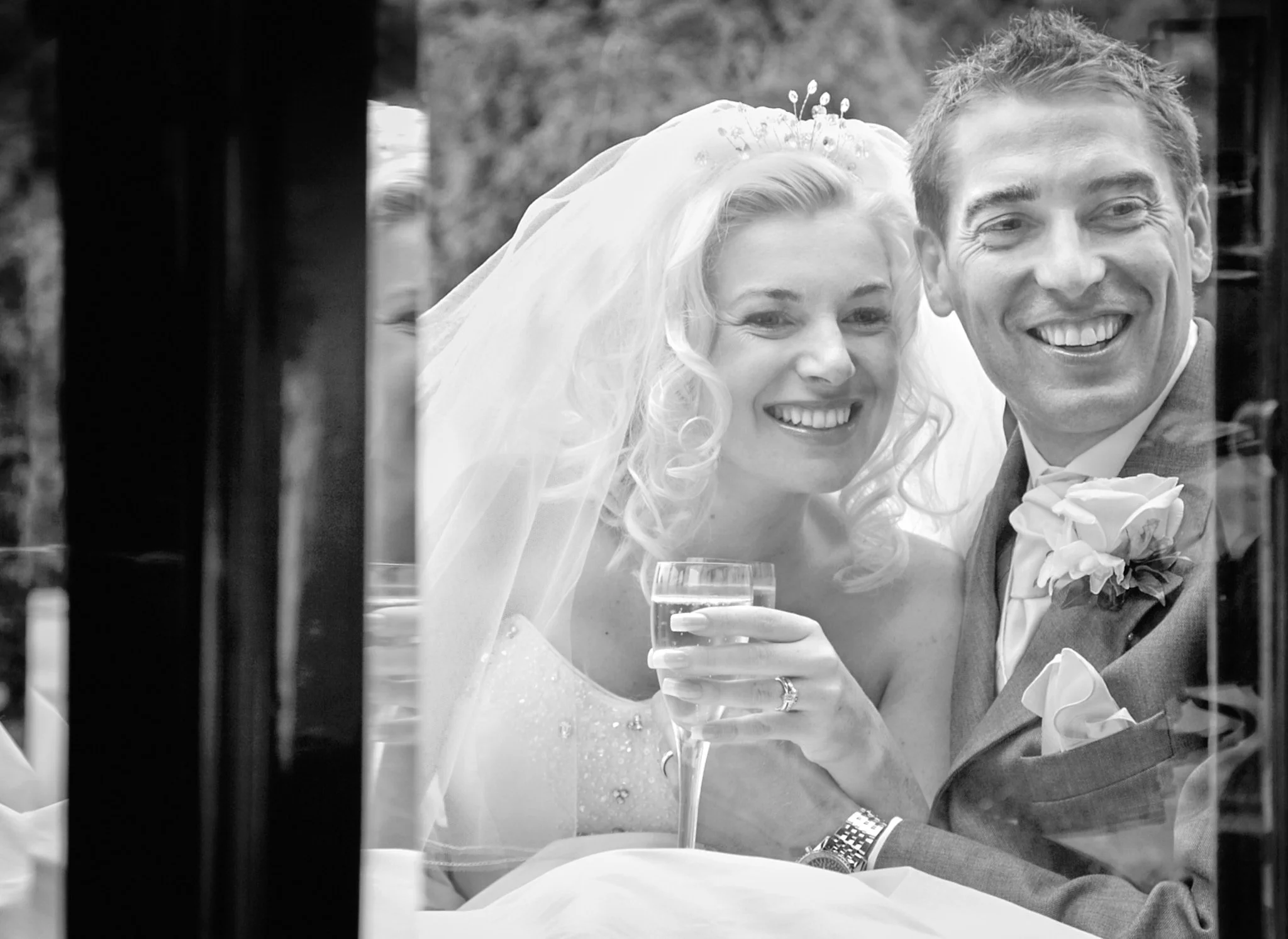 Black and white photo of a smiling bride and groom at their wedding, seen through a window or partition.