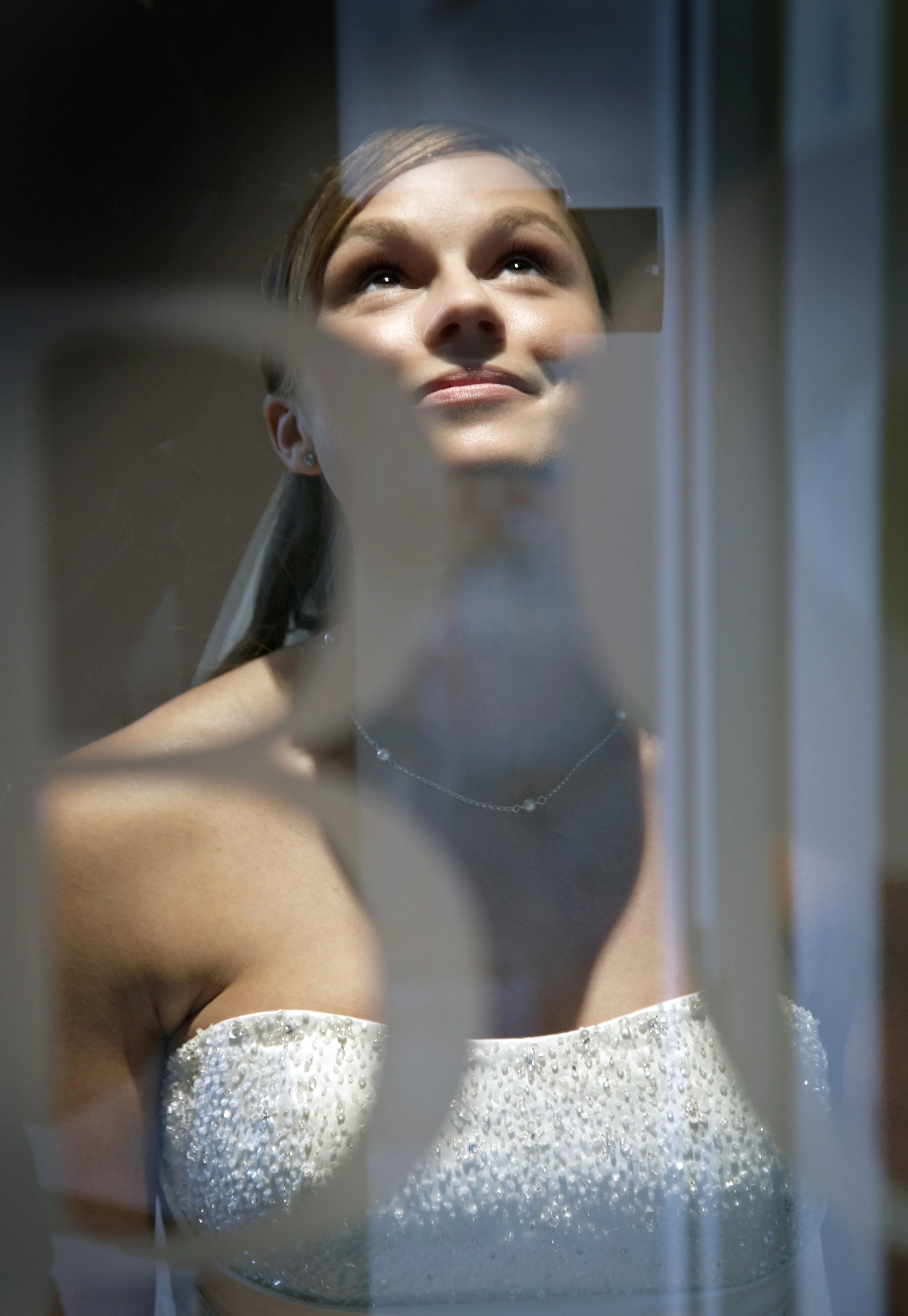 Young woman in wedding dress looking up, reflected on mirror with frosted glass in foreground.