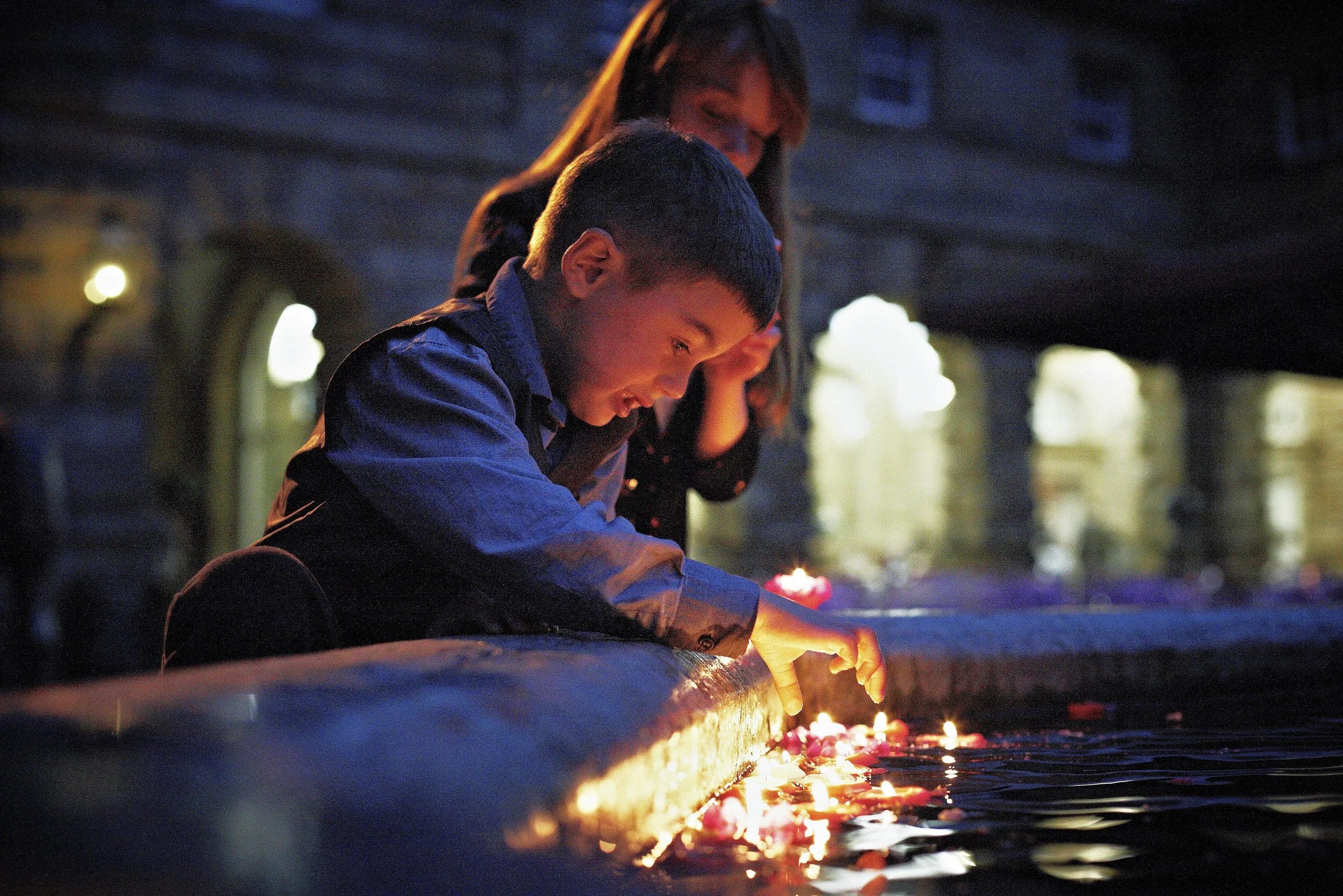 A young boy and a woman lighting candles on a ritual or religious ceremony at dusk, with a background of a building and faint lights.