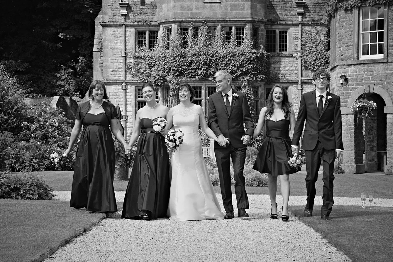 Black and white photograph of a wedding party walking on a gravel path outside a stone building, holding hands and smiling, including the bride in a white gown, the groom in a dark suit, and four bridesmaids and groomsmen in formal attire.