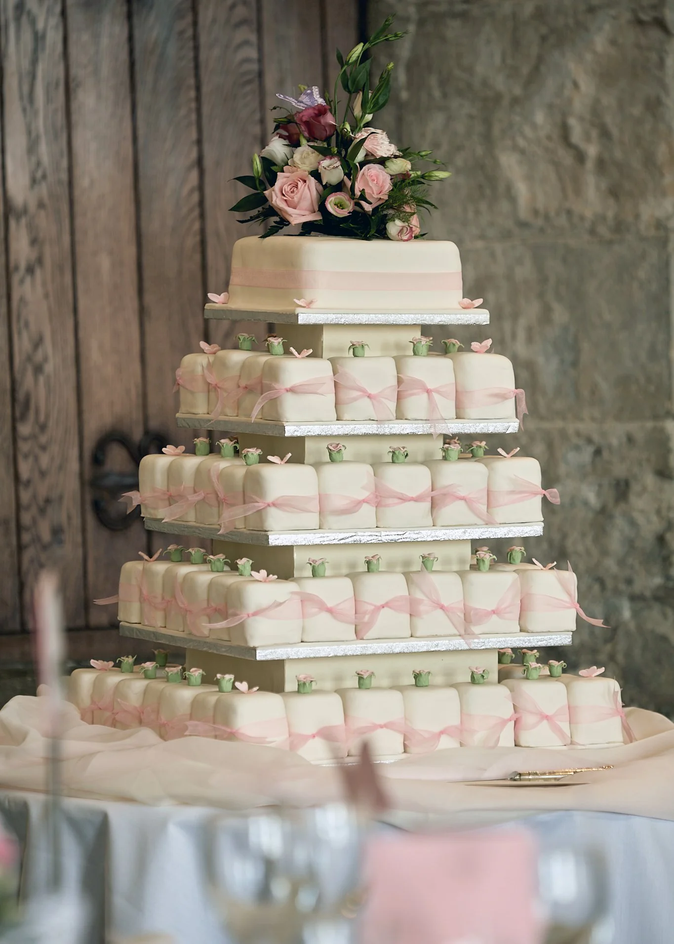 Tiered wedding cake decorated with pink ribbons, small green and pink cupcake toppers, topped with a bouquet of pink and white roses, greenery, and butterflies.