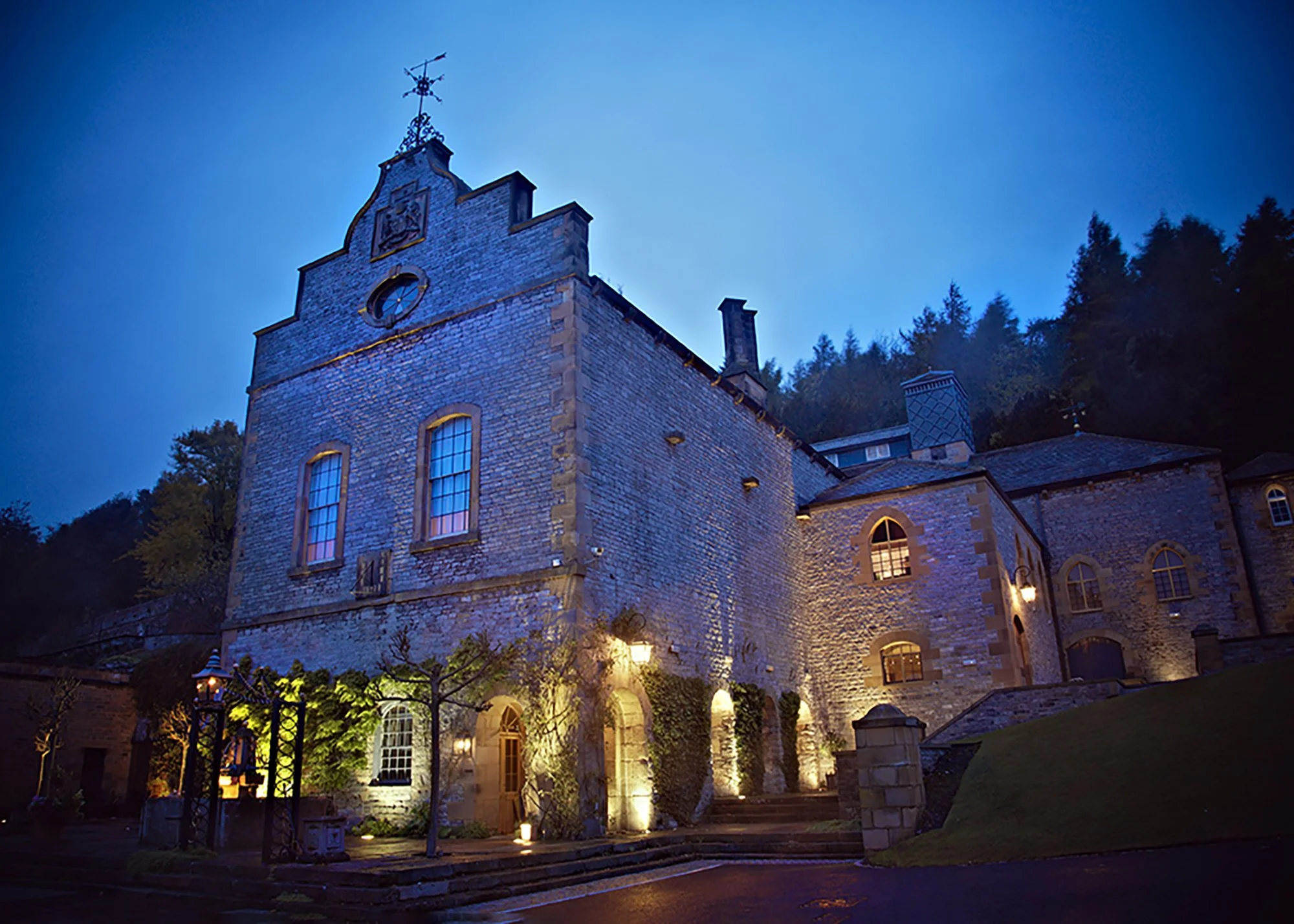 A large stone building with tall, arched windows illuminated from inside, surrounded by landscaped garden and lit with exterior lights at dusk or night.