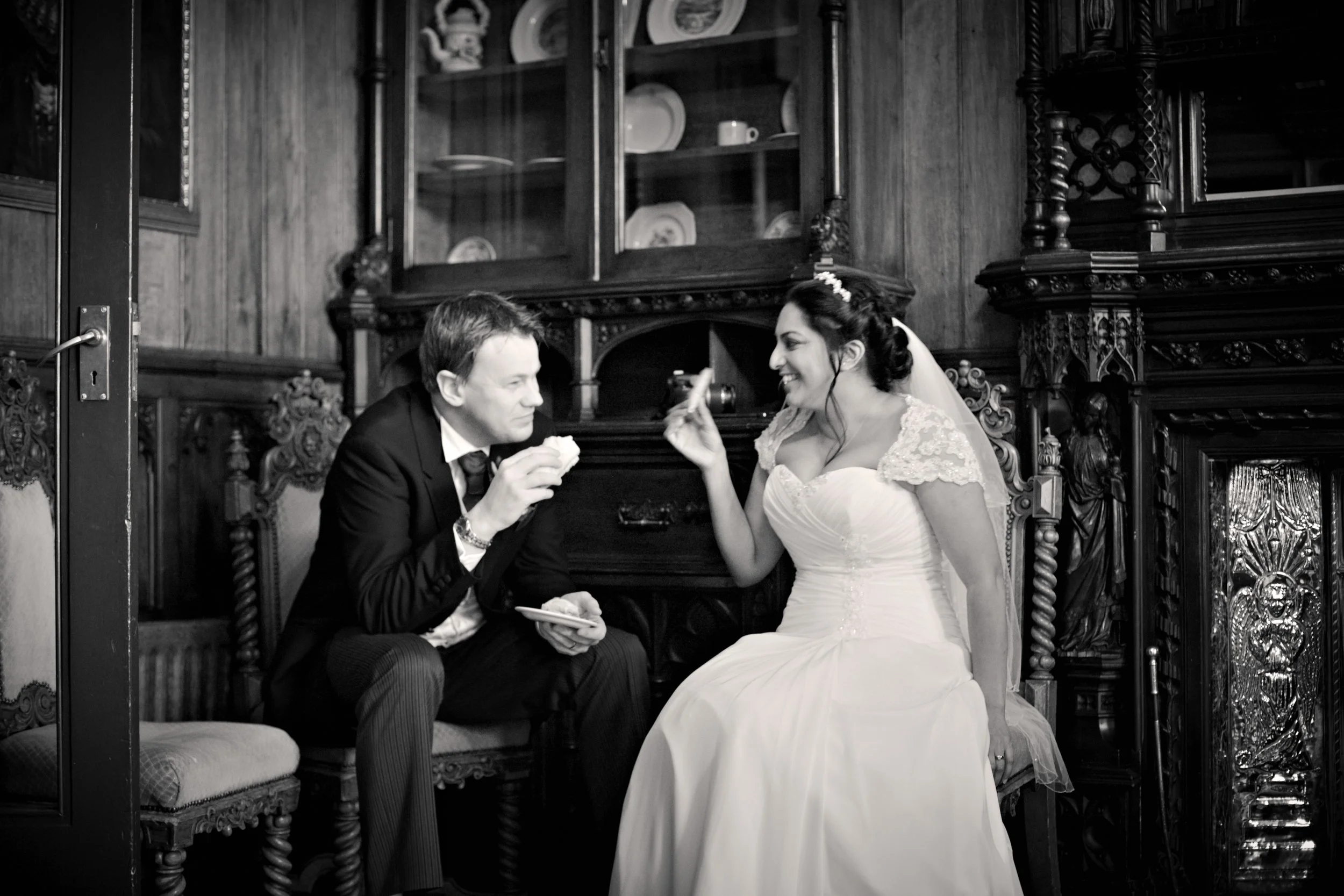 A bride and groom sitting on a carved wooden bench in a vintage-style room, smiling and sharing a moment, with the groom offering a piece of cake to the bride.