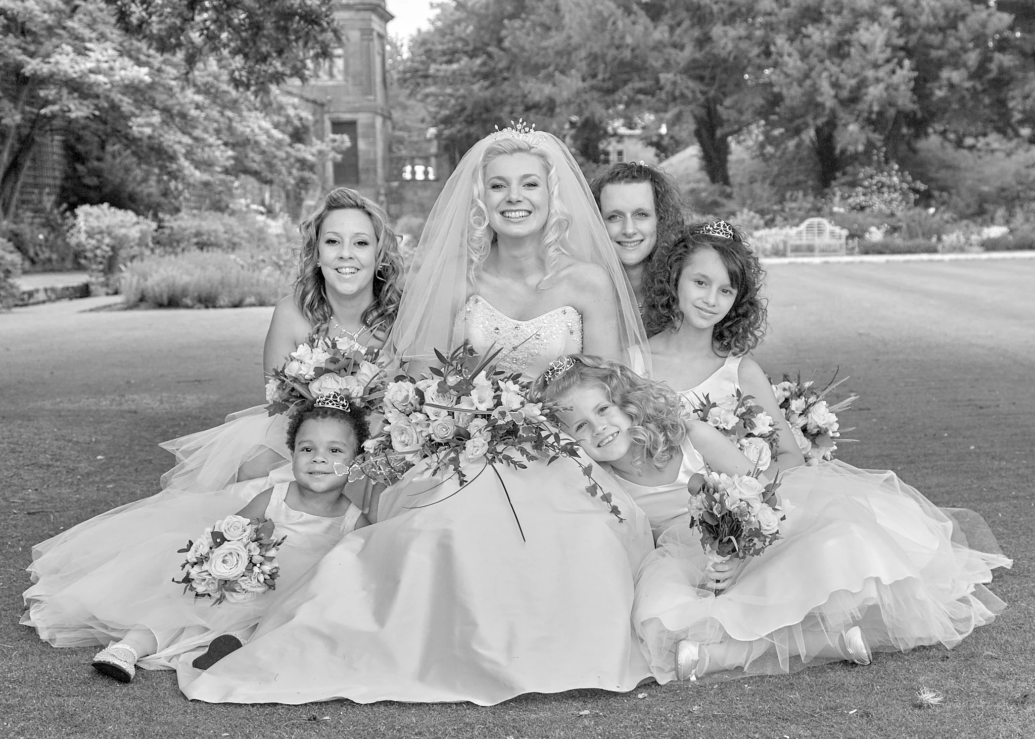 Black and white photo of a bride with curly blonde hair wearing a wedding dress and veil, surrounded by young girls dressed as princesses with tiaras, all holding bouquets of flowers, in an outdoor garden setting.