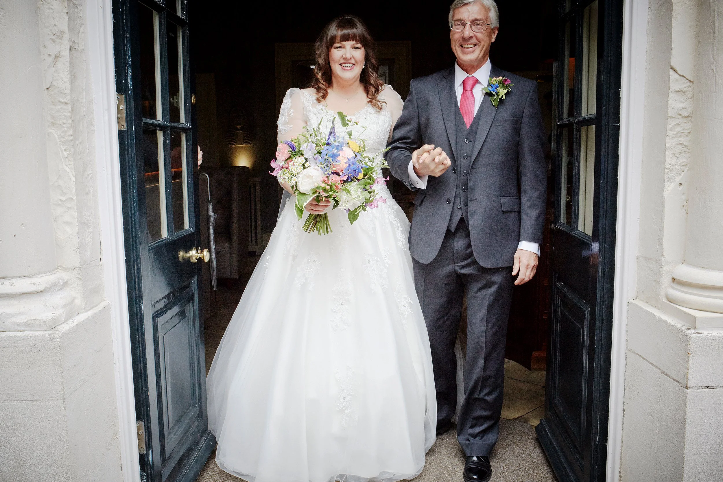A bride and an older man, possibly her father, standing at a doorway, smiling. The bride is holding a colorful bouquet and wearing a white wedding gown with lace details. The man is dressed in a dark suit with a vest, white shirt, pink tie, and has a