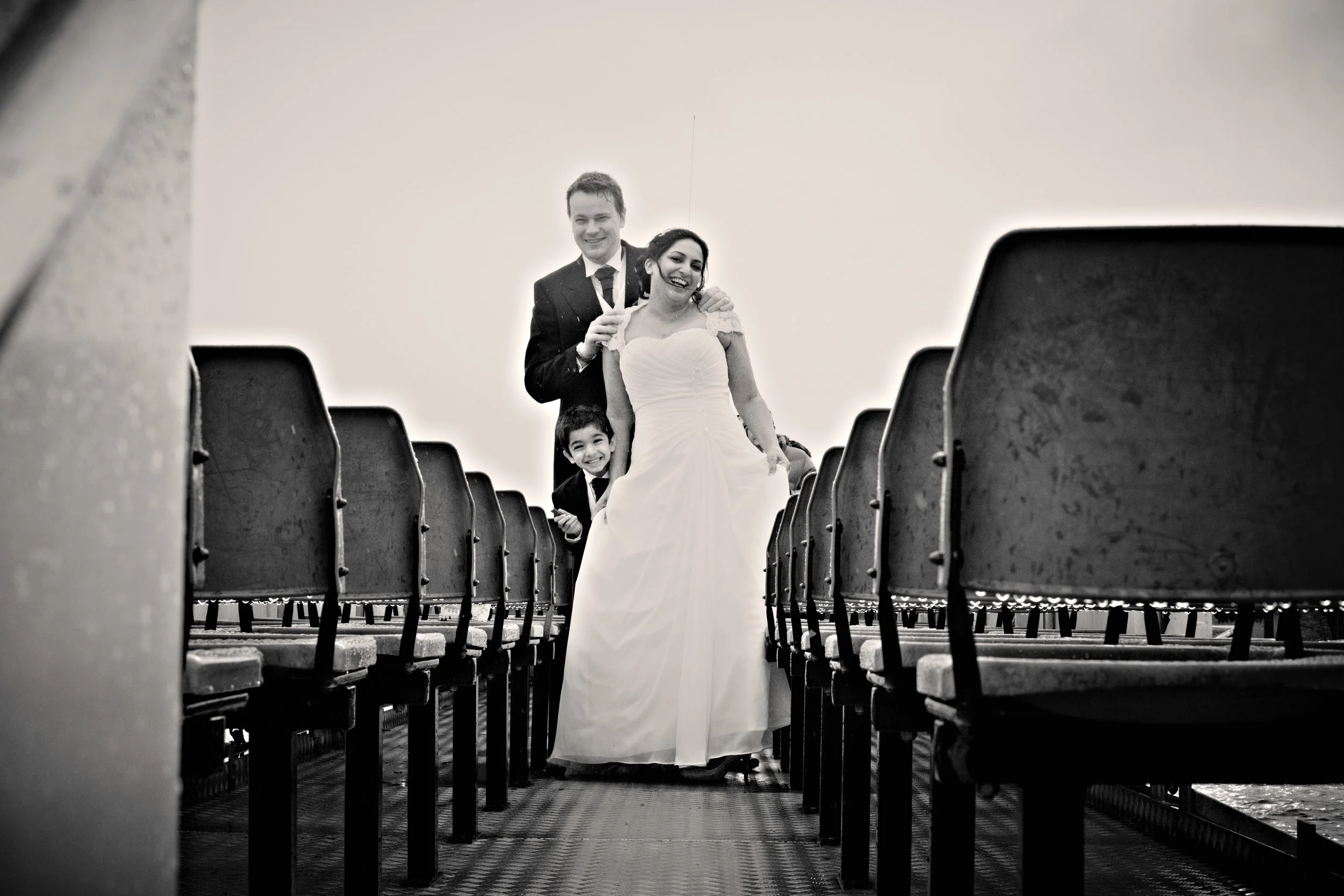 A wedding couple, a groom and a bride, walking down a staircase with a young girl, all smiling, in an outdoor setting with empty stadium seating around them, in black and white.
