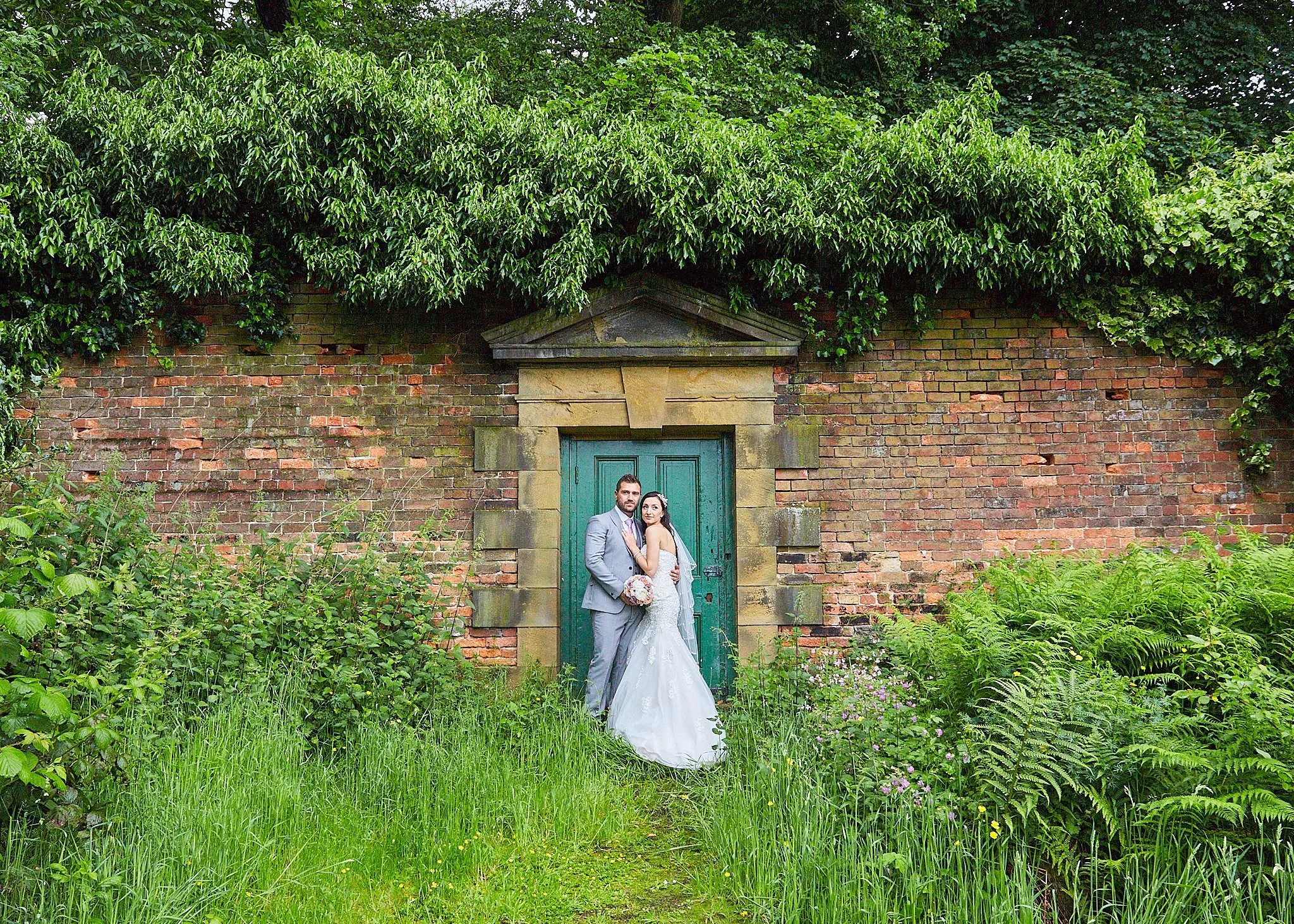 A bride and groom in wedding attire standing close together in front of an old brick wall with a green door, surrounded by lush overgrown greenery and bushes.