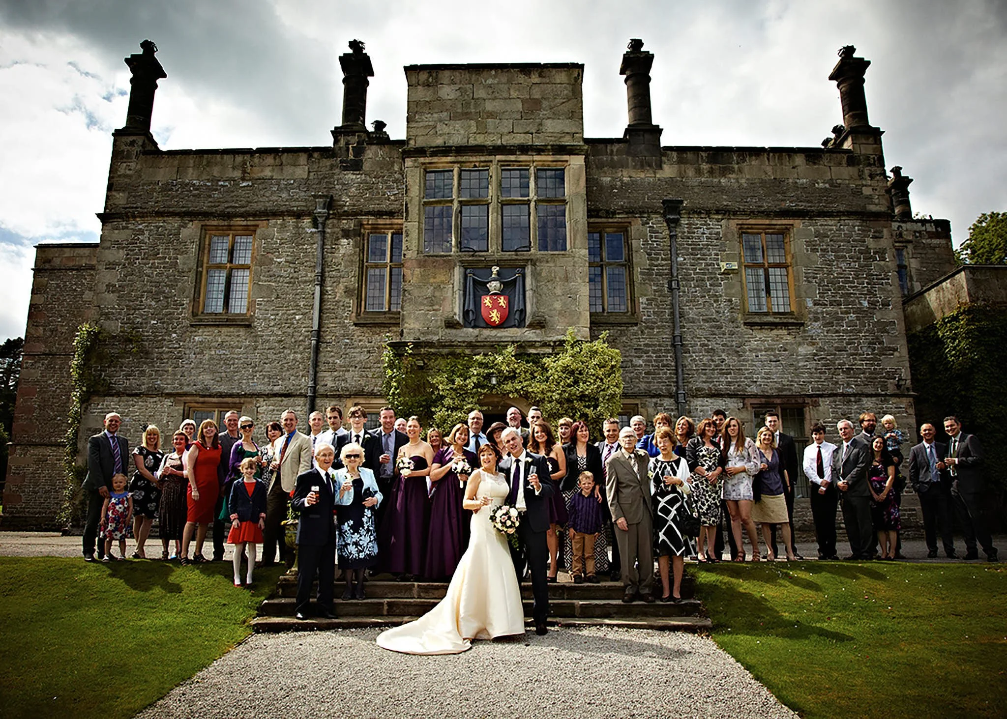 A large group of people standing on steps and grass in front of a historic stone building with tall chimneys, during a wedding celebration. The bride and groom are in the front, with the bride in a white gown holding a bouquet and the groom in a dark