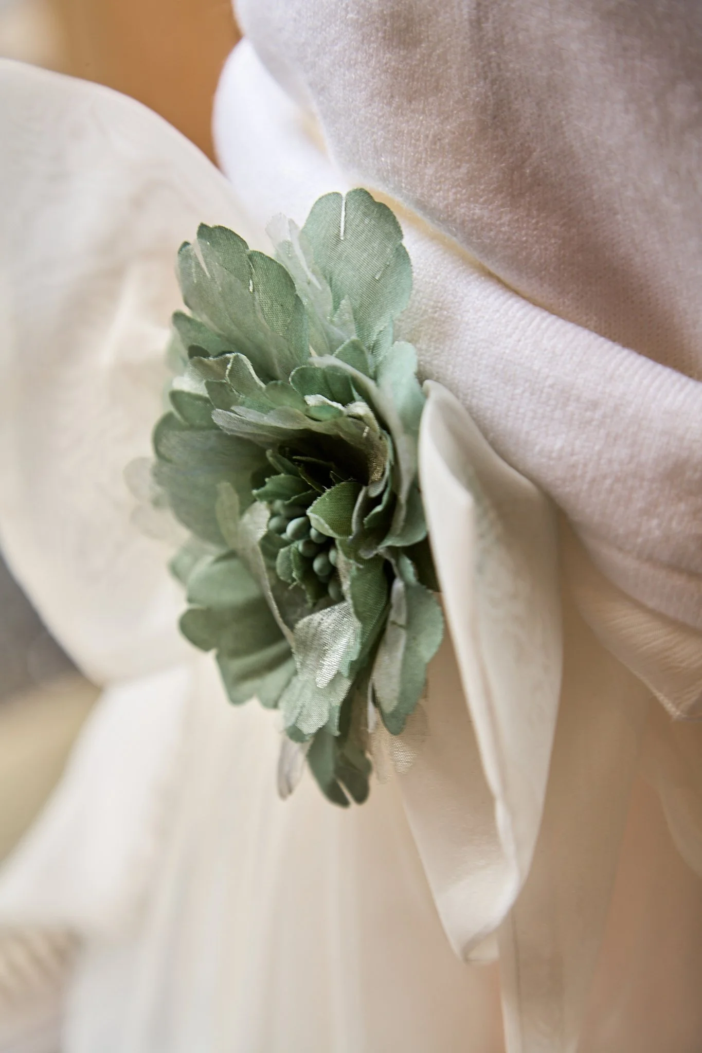 Close-up of a boutonniere with green leaves attached to a cream-colored fabric, likely part of a wedding dress or suit.