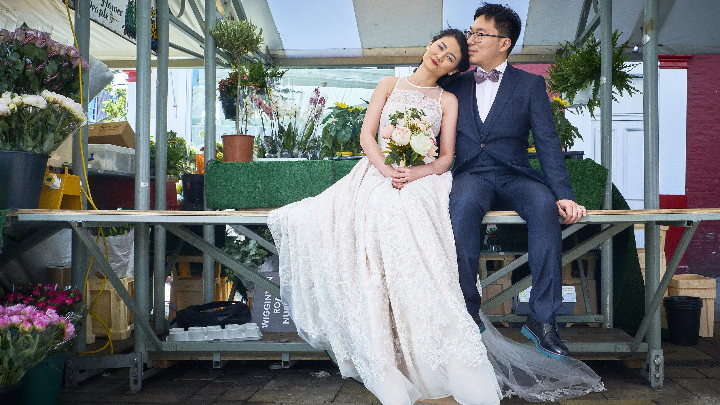 A bride and groom sitting on a stage at a florist shop, with flowers around them. The bride is in a white wedding dress holding a bouquet, and the groom is in a dark suit with glasses and a bow tie.