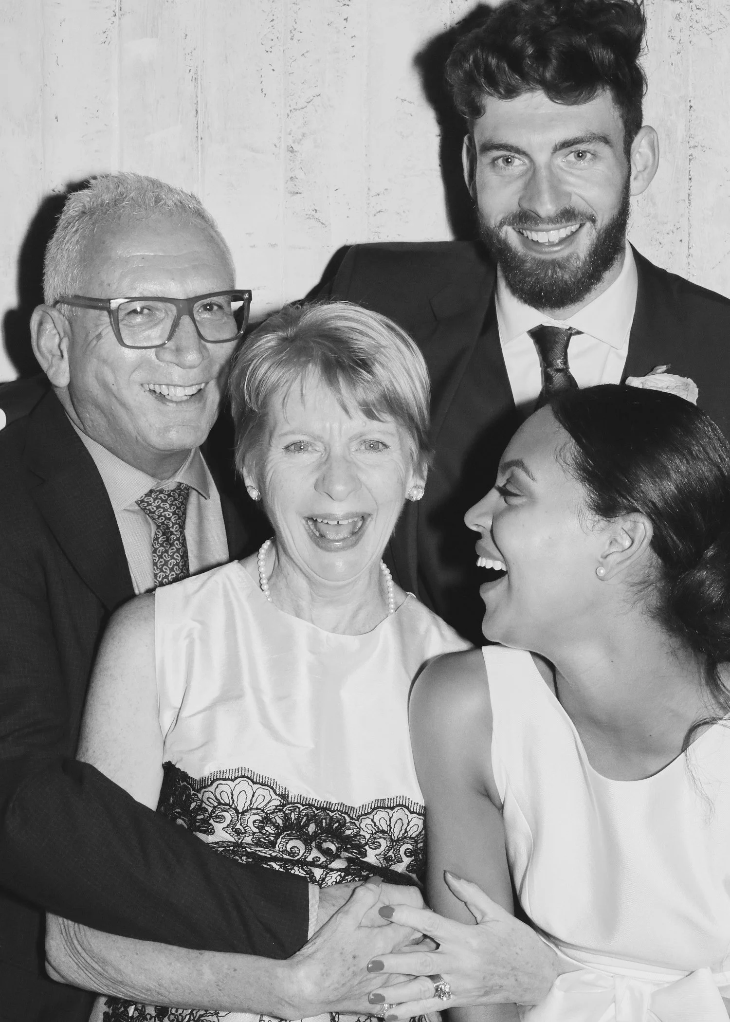 Four people smiling and laughing, two women and two men, in a close group. Their hands are held together in the center of the image. The background appears to be a plain wall.