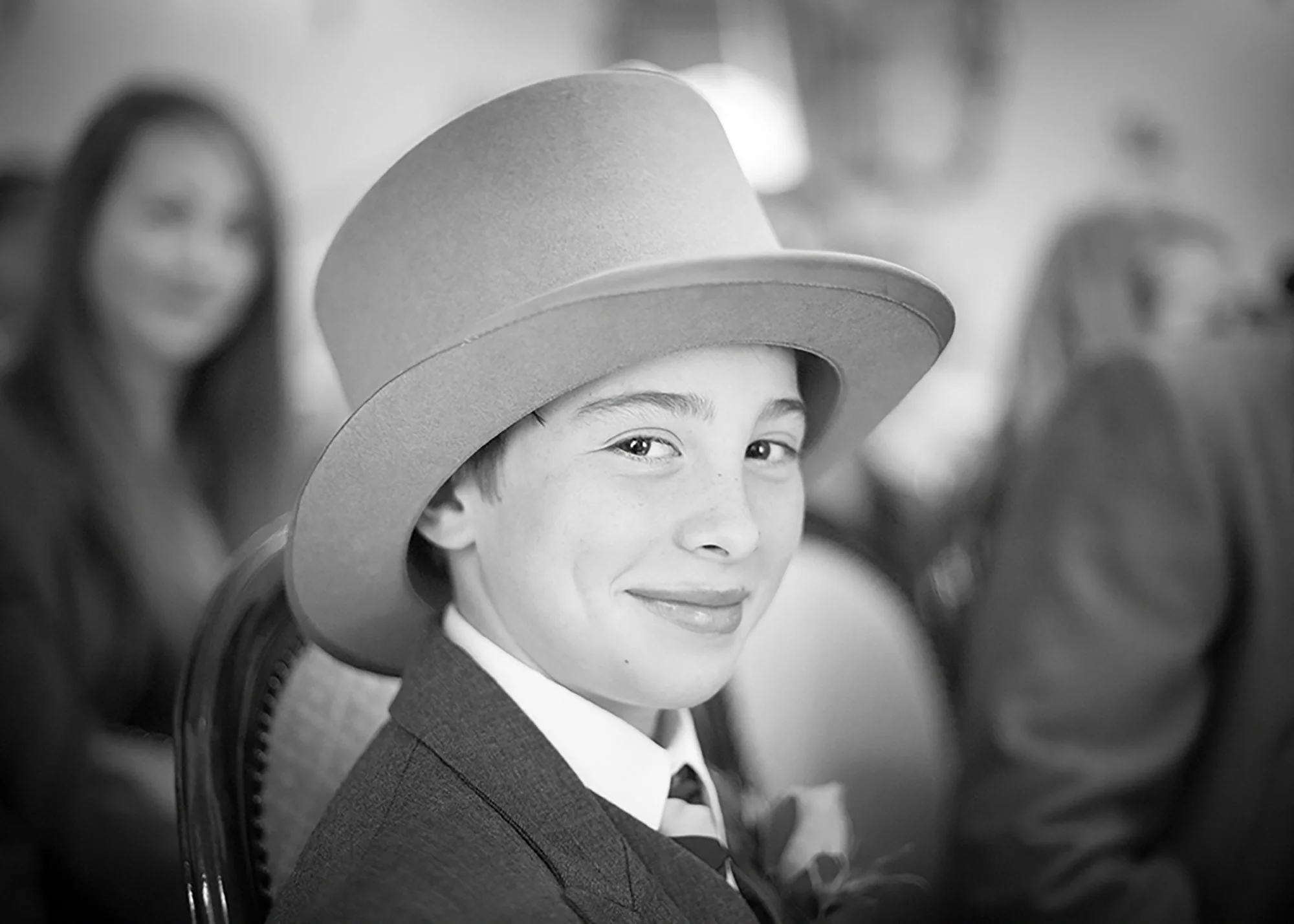 A young boy dressed in formal attire, wearing a hat, sitting on a decorative bench, smiling and looking at the camera, with blurred women in the background.