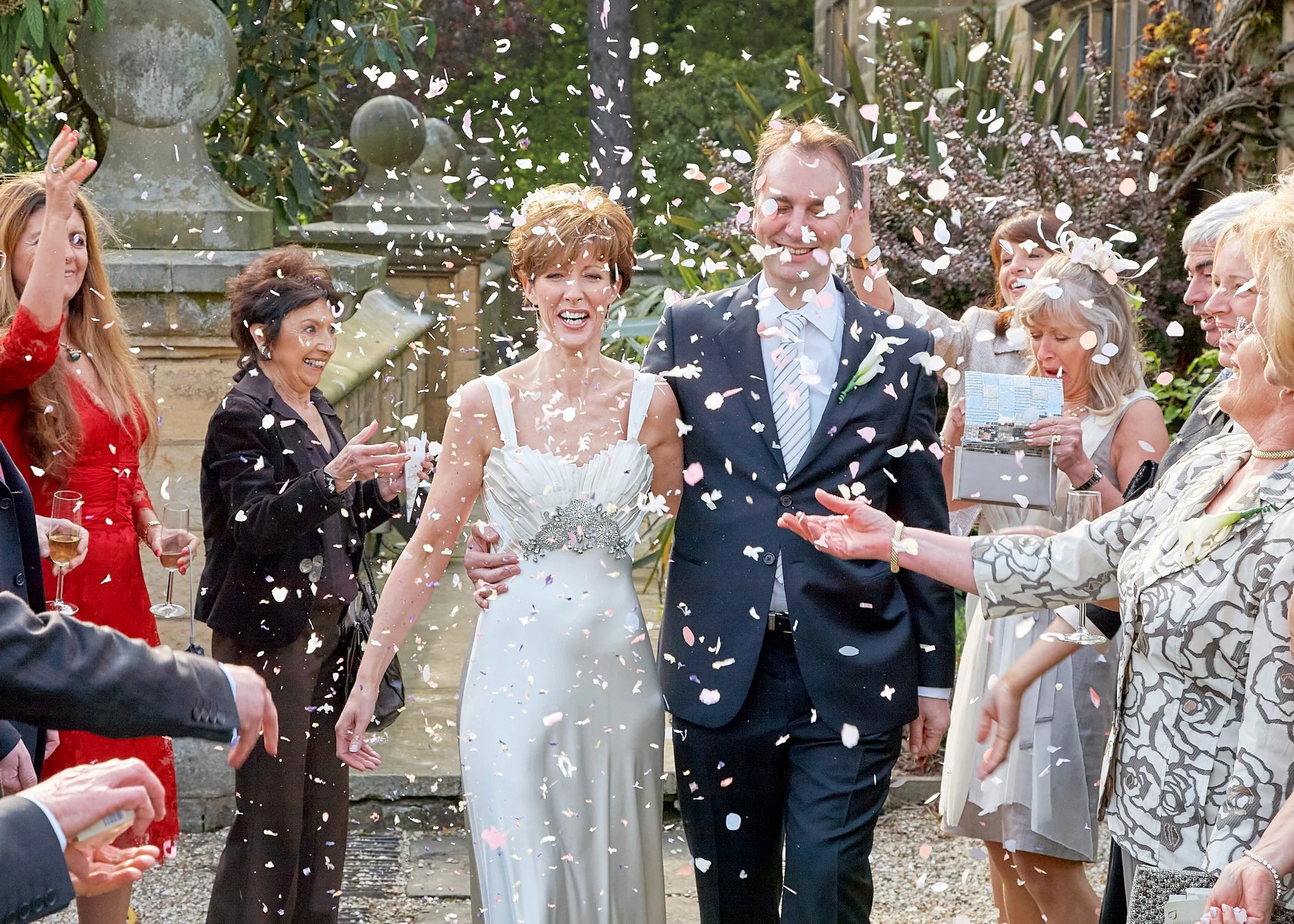 A wedding celebration with people throwing flower petals into the air as a bride and groom smile. The scene is outdoors with greenery and stone statues in the background.