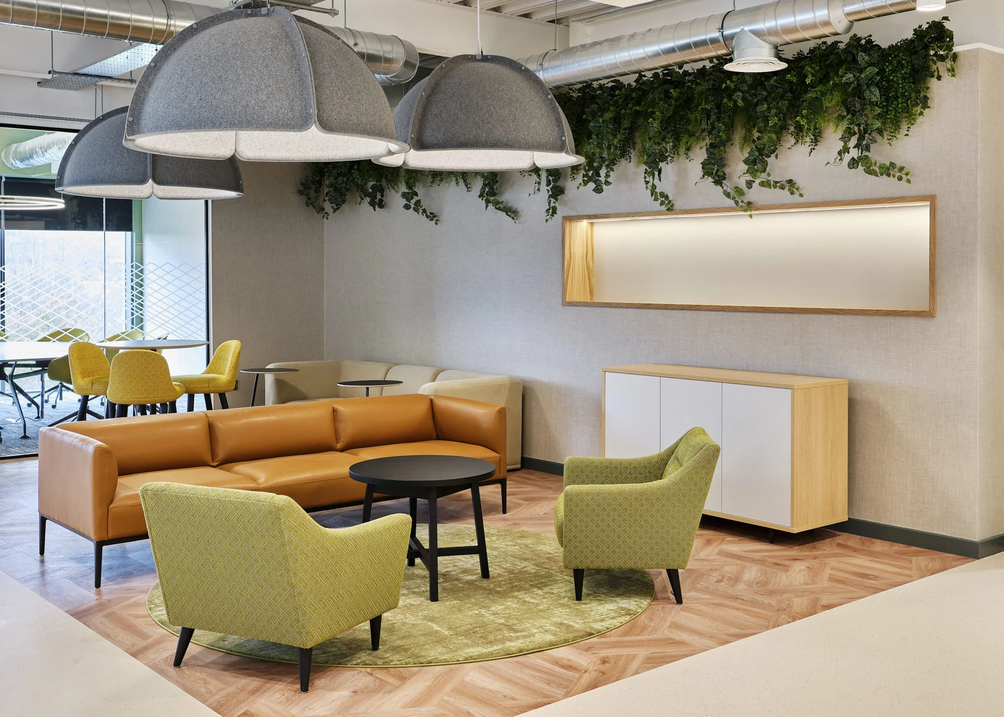 Modern interior lounge area with orange sofa, green armchairs, black coffee table, hanging grey felt lamps, and green plants on the wall.