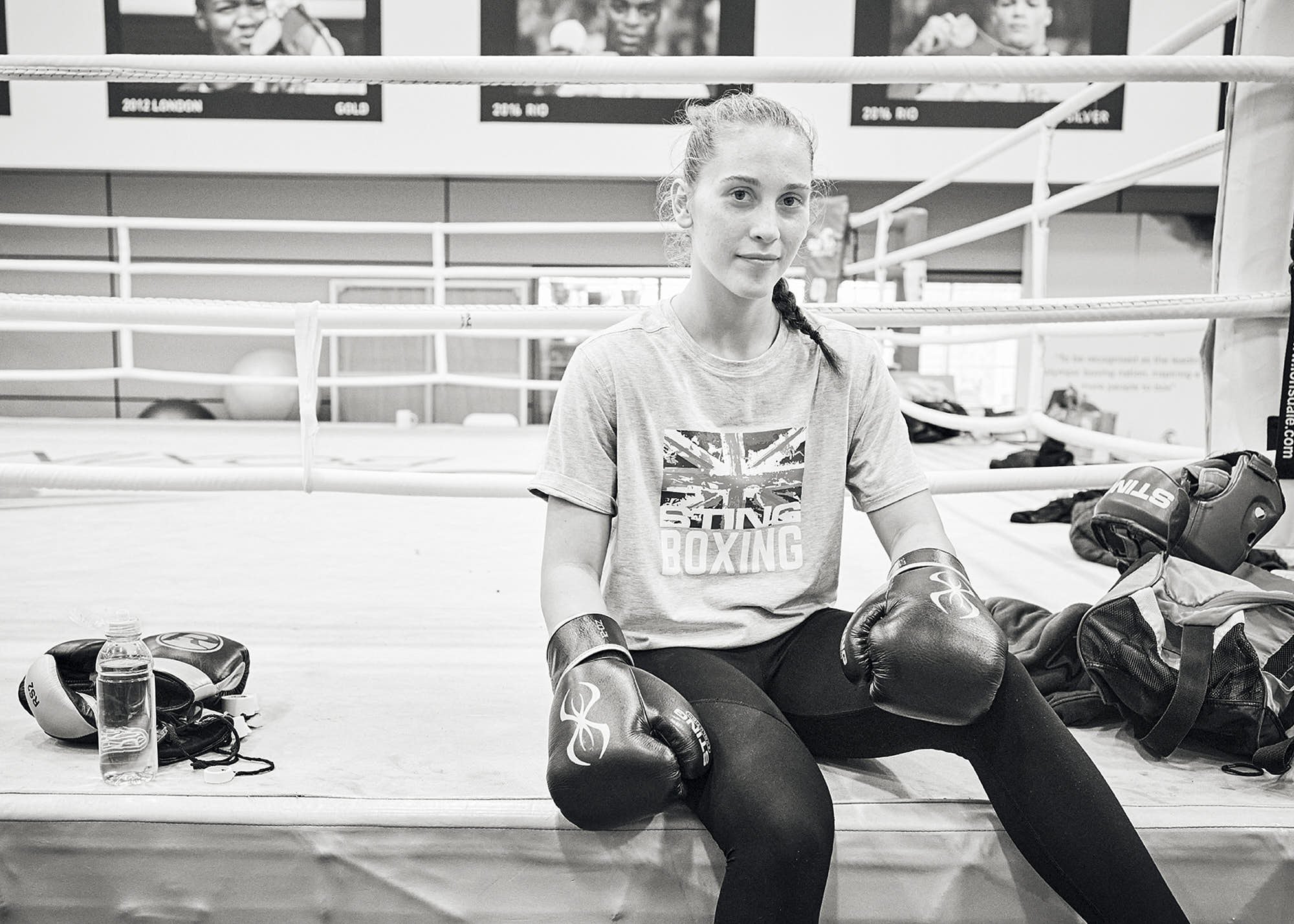 A young woman sitting inside a boxing gym, wearing boxing gloves, with boxing equipment and a water bottle nearby. The gym has boxing rings and motivational posters on the wall.