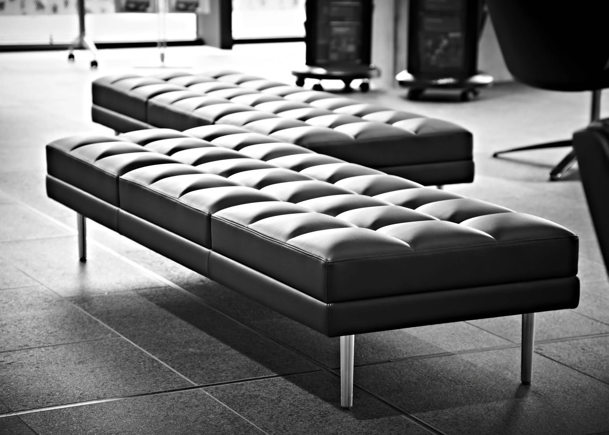 Black leather tufted bench with metal legs in an office or waiting area, with windows and other furniture visible in the background.
