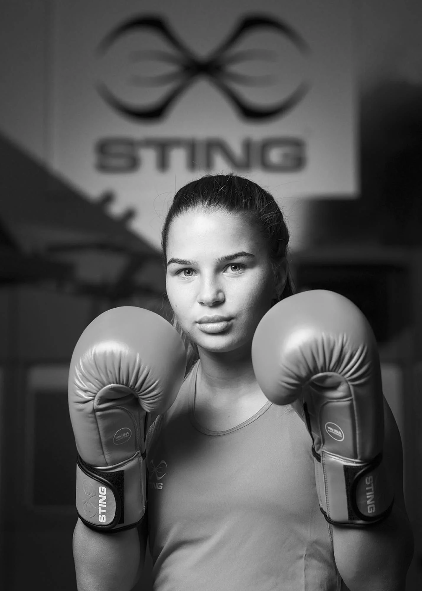 Black and white photo of a young woman in boxing gloves, positioned in a fighting stance with a serious expression, in front of a wall with a sign that reads "STING" and a stylized logo.