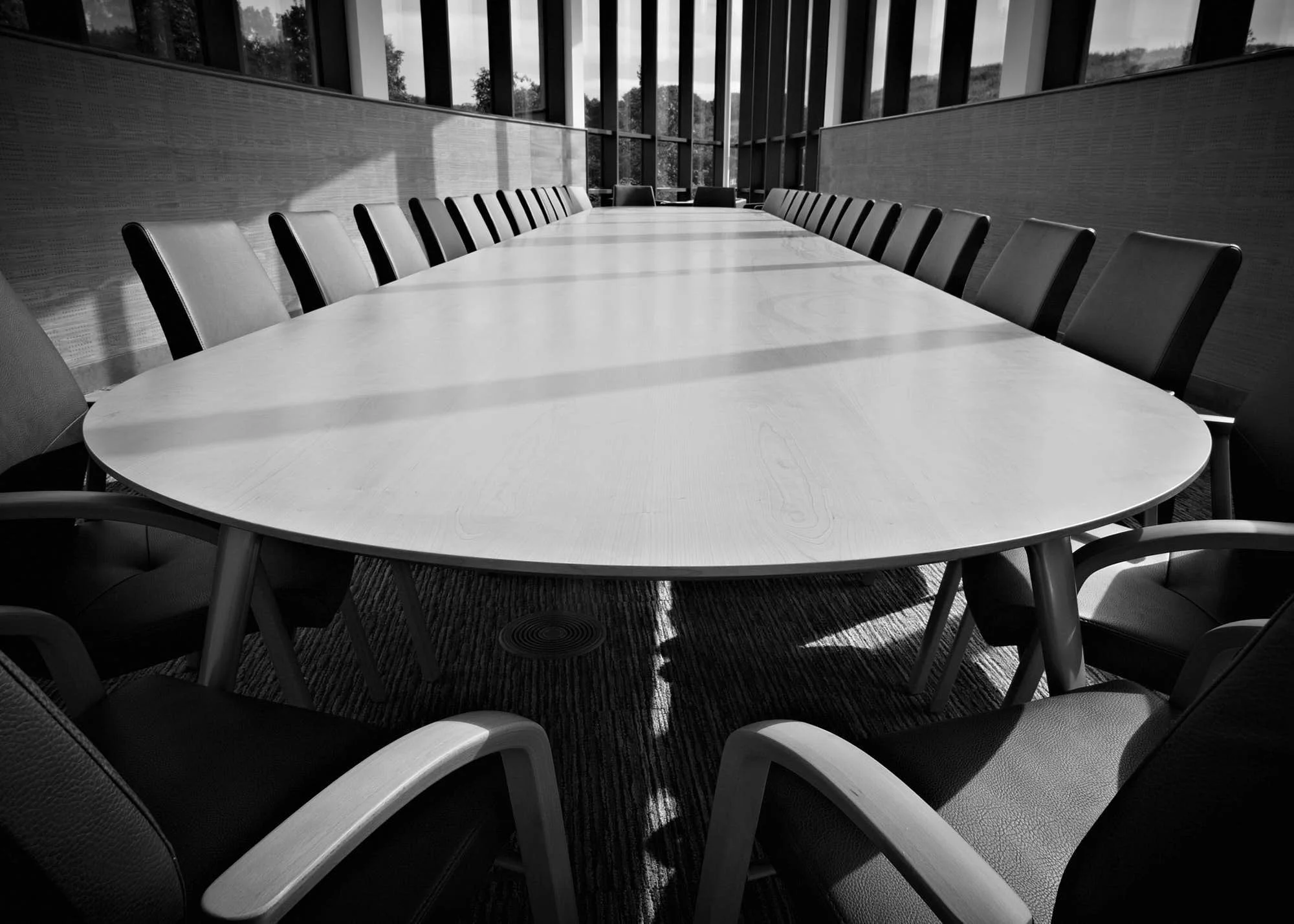A large conference room with a long oval table surrounded by leather chairs, large windows with vertical blinds, and a view of trees outside.