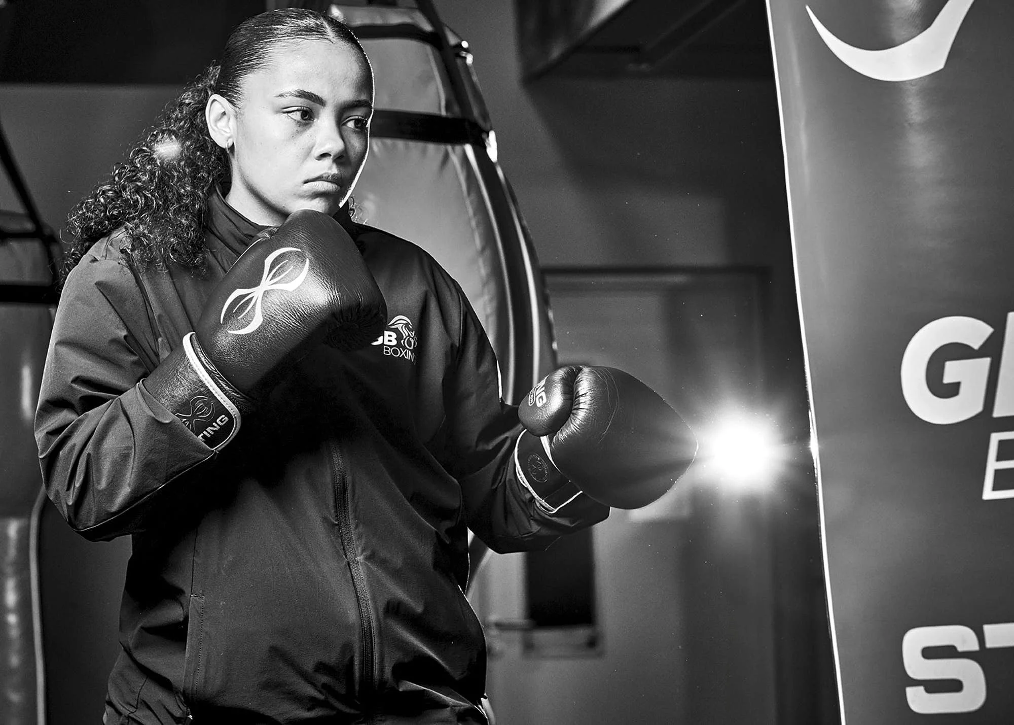 A woman in boxing gear, including gloves and a jacket with a logo, stands in front of a banner in a boxing gym.