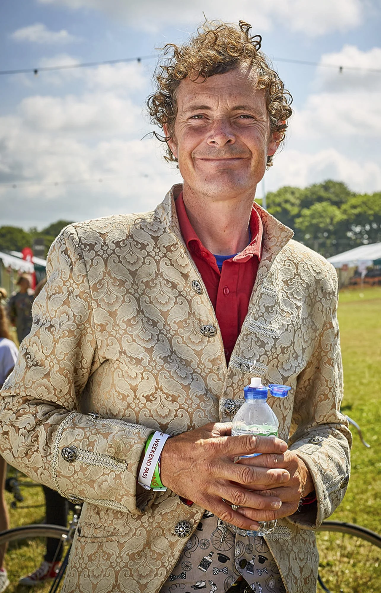 A man with curly hair and a beige patterned blazer holding a water bottle, smiling outdoors on a sunny day.