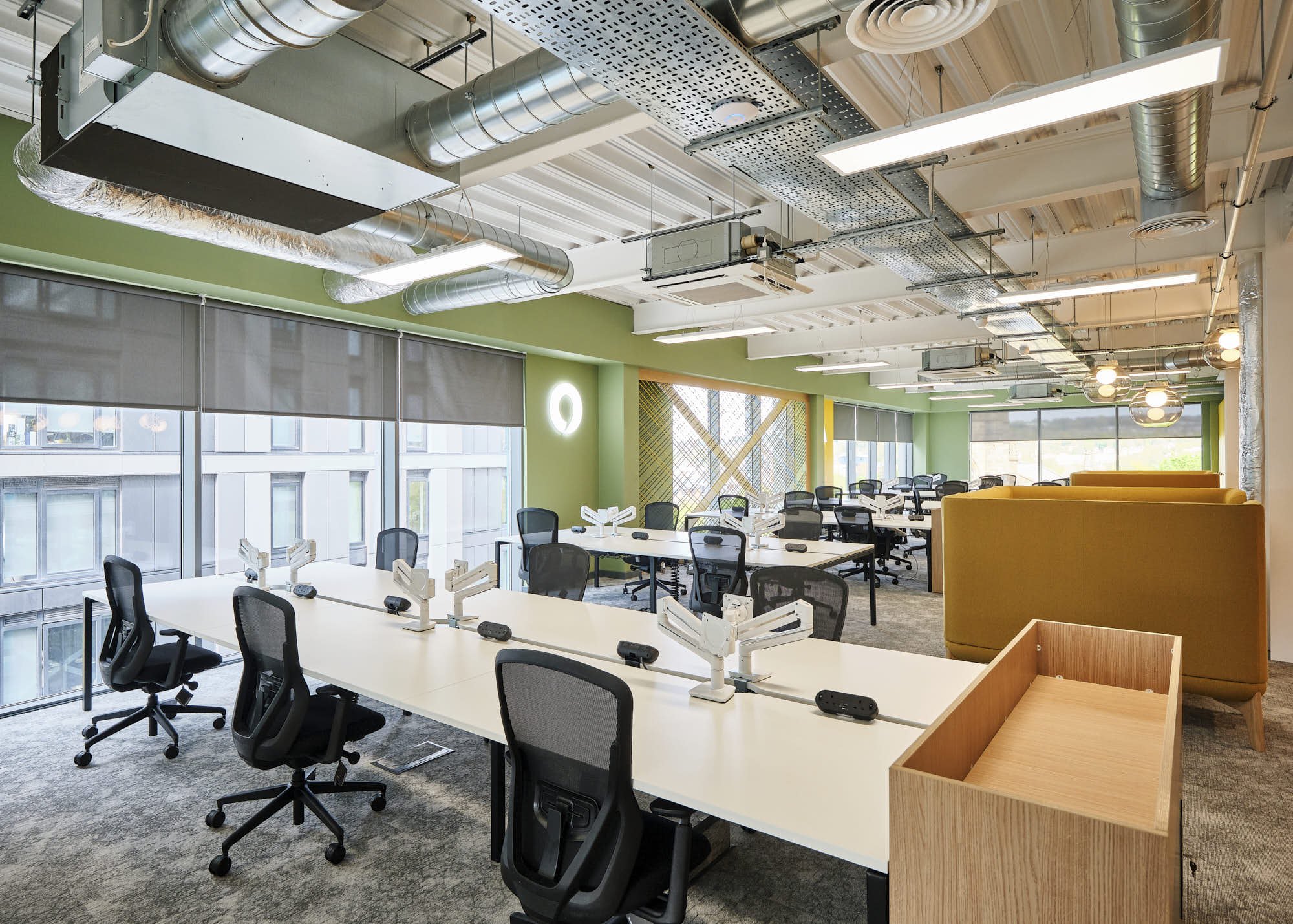 Modern office space with white desks, black ergonomic chairs, pendant lighting, large windows with gray shades, exposed ductwork on ceiling, and a green accent wall.