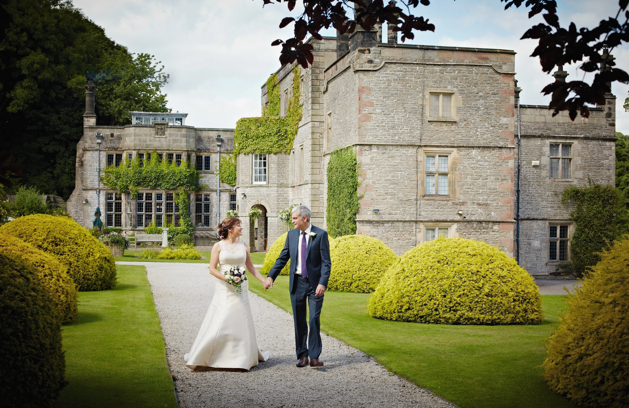 A bride and groom walking hand-in-hand along a stone pathway in front of an old stone mansion with ivy-covered walls, surrounded by lush green trees and bushes.