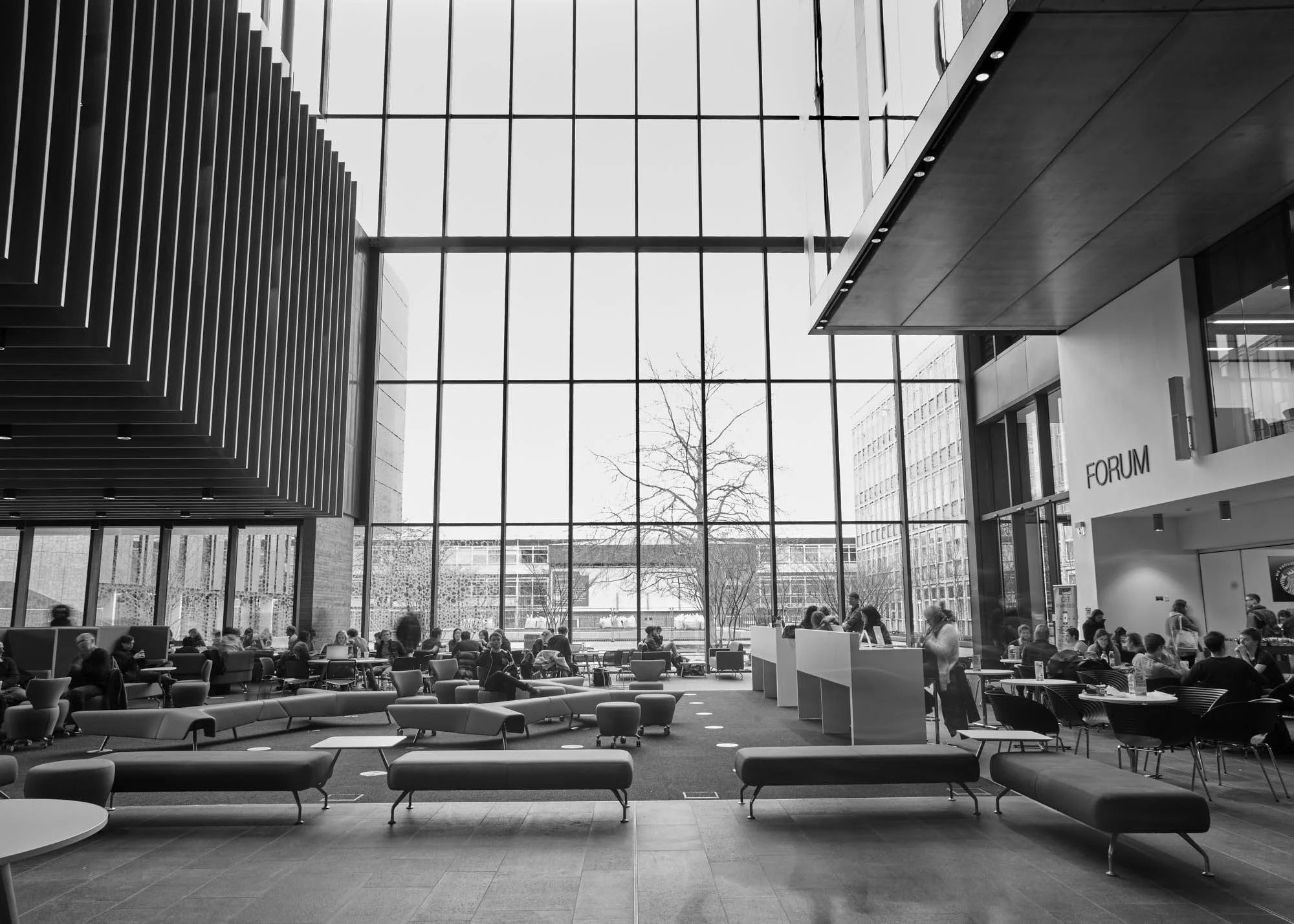 Interior view of a modern, spacious hall with large glass windows letting in natural light. There is a seating area with several chairs and tables, and people sitting and working. A sign reads 'Forum' on the right side of the image.