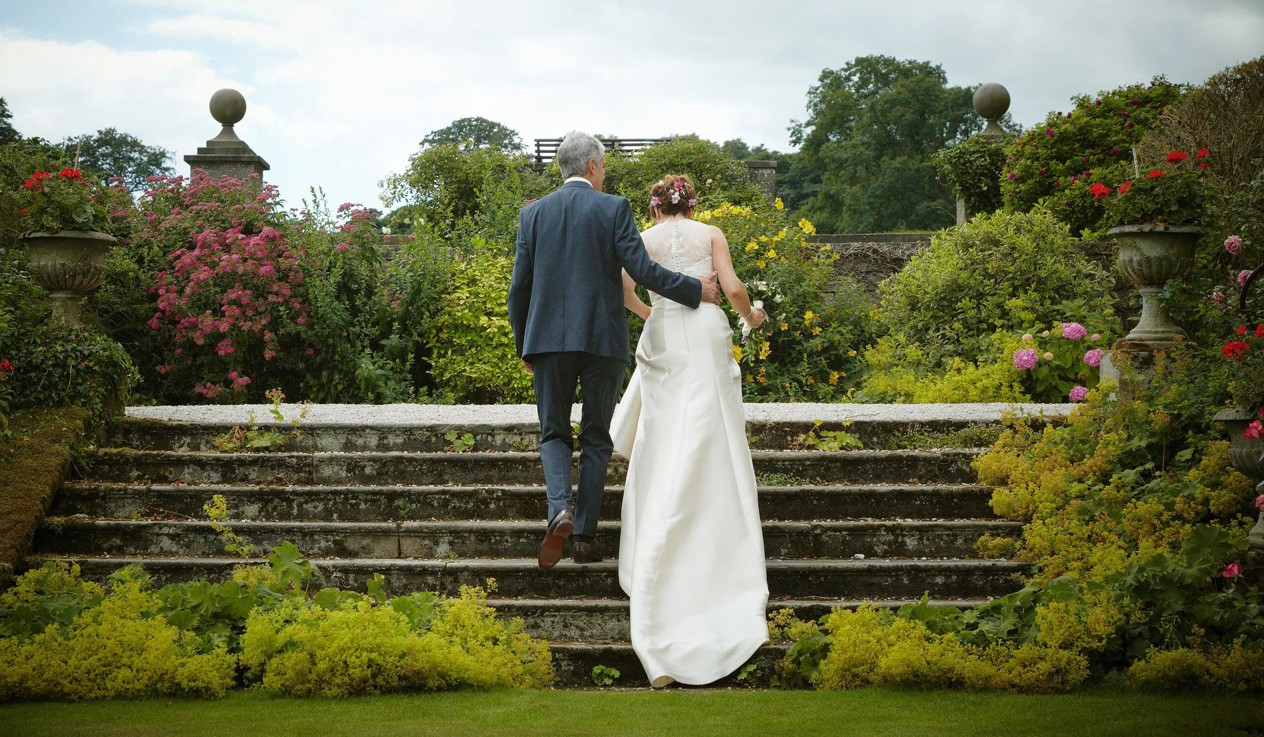A couple dressed in wedding attire walking up stone steps in a lush garden, with the woman holding a bouquet and wearing a flower crown.