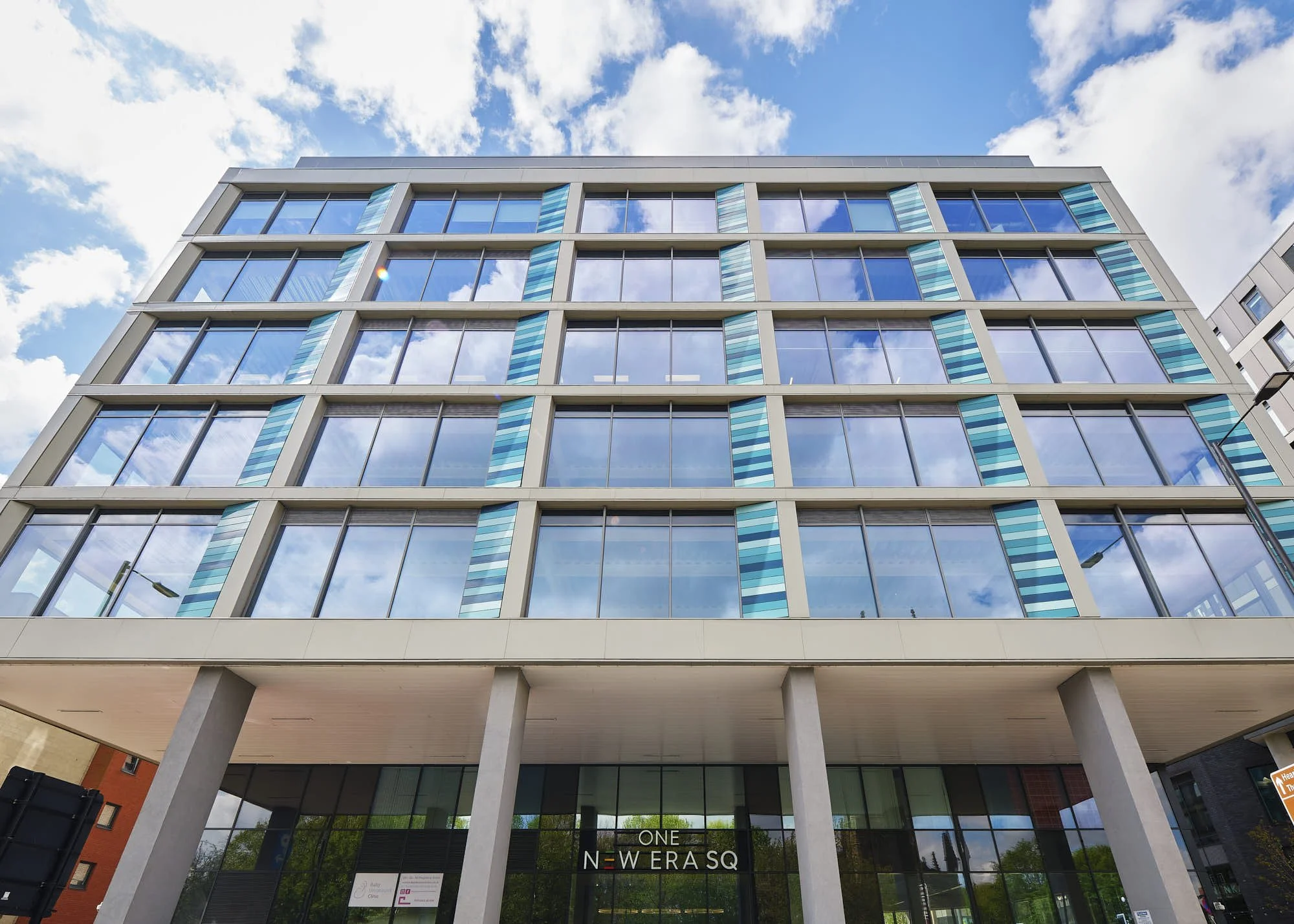 Modern multi-story office building with glass windows reflecting the sky, labeled 'ONE N E WA ER SQ' at the entrance, with trees visible behind.