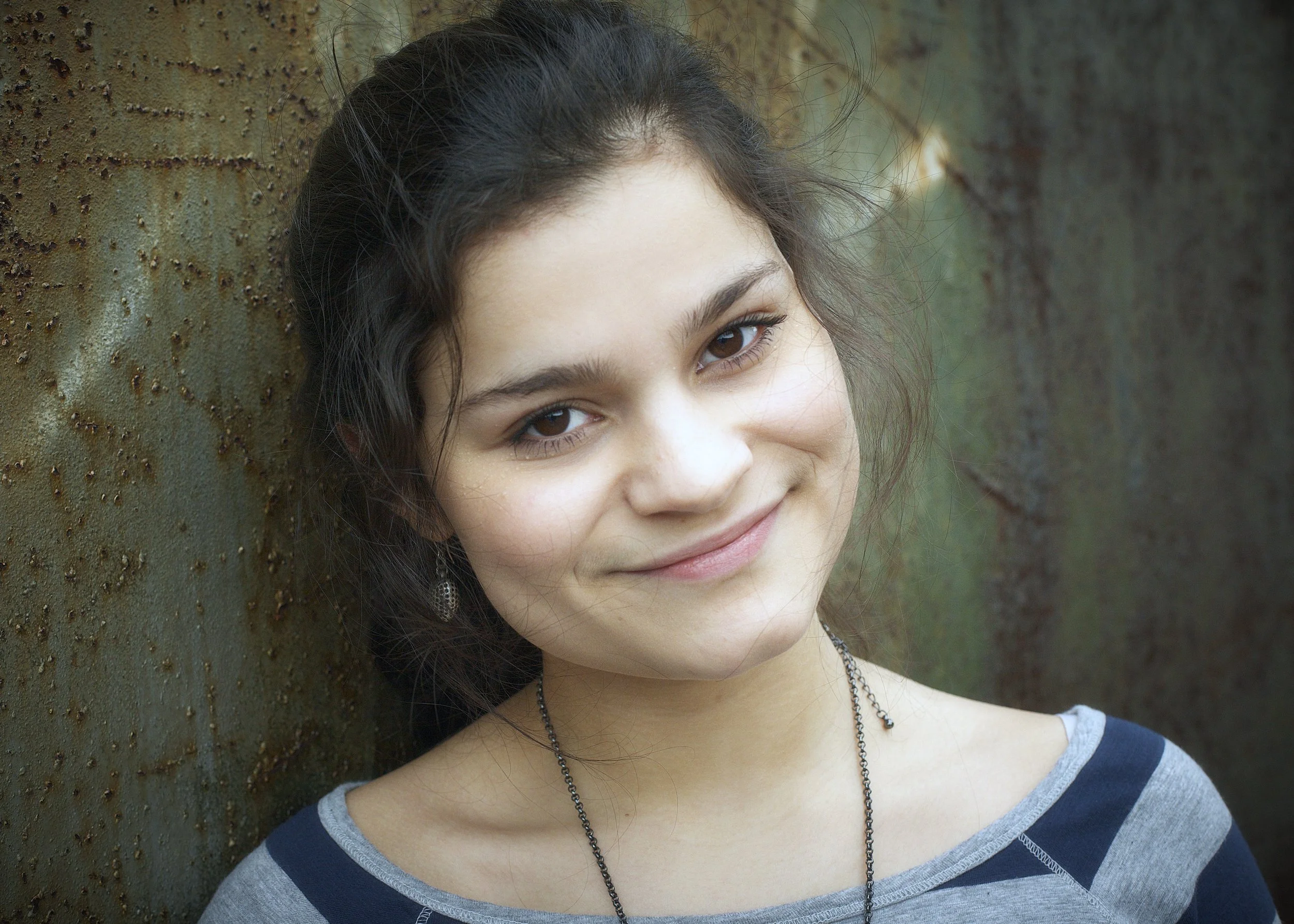 Close-up of a young woman smiling with brown eyes and dark hair, wearing a striped shirt and a necklace, leaning against a textured, rusty, metal surface.