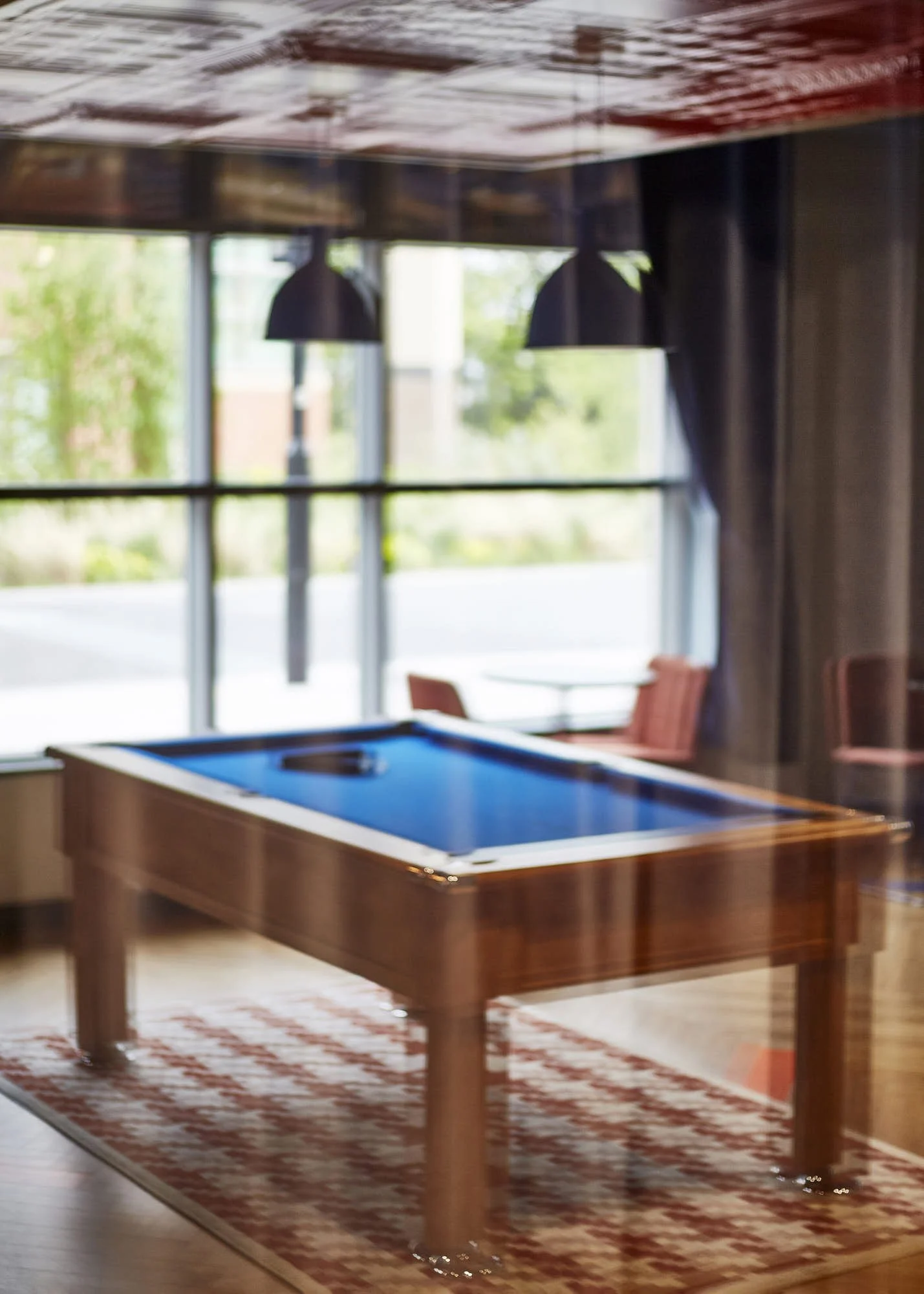 Indoor scene with a pool table on a red patterned rug, black pendant lights hanging from the ceiling, large windows with a view of green trees outside, and wooden chairs in the background.