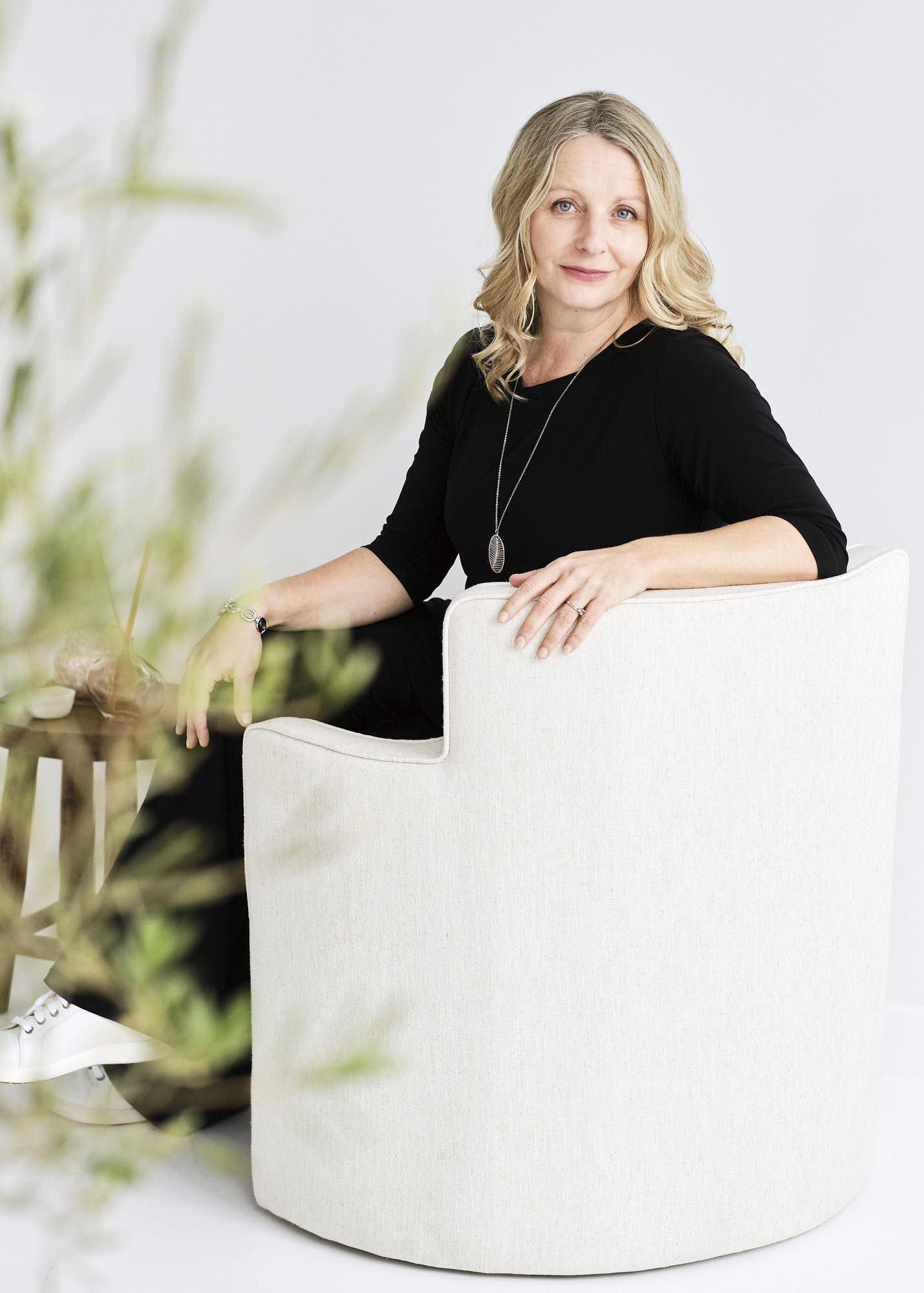 A woman with blonde hair wearing a black top and a necklace seated on a white chair, with blurred foreground greenery and a plain white background.