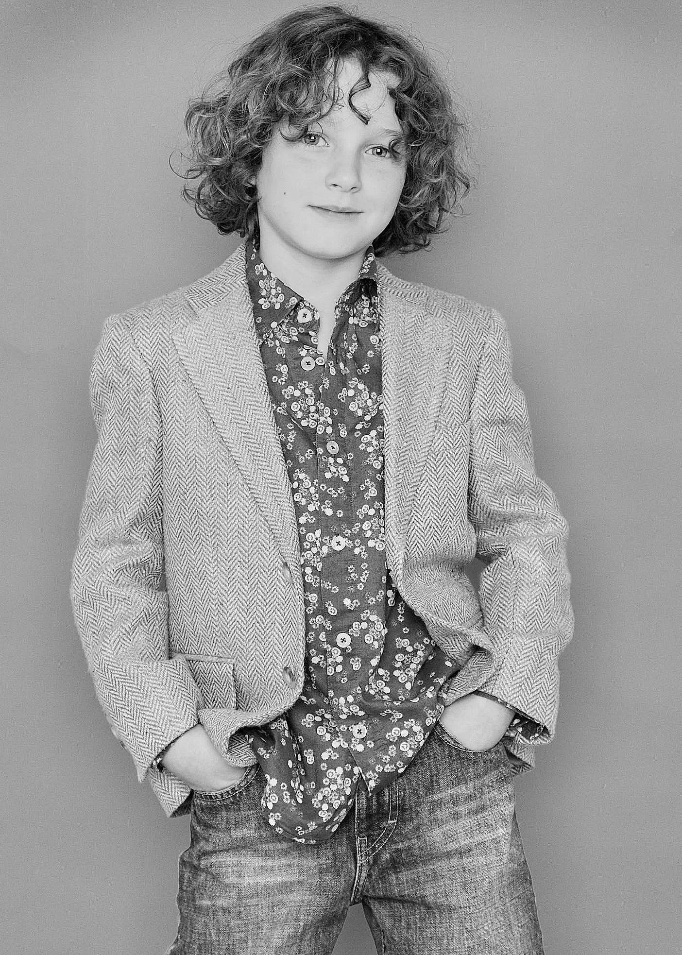 Black and white photo of a person with curly hair, wearing a patterned shirt, blazer, and jeans, standing against a plain background.
