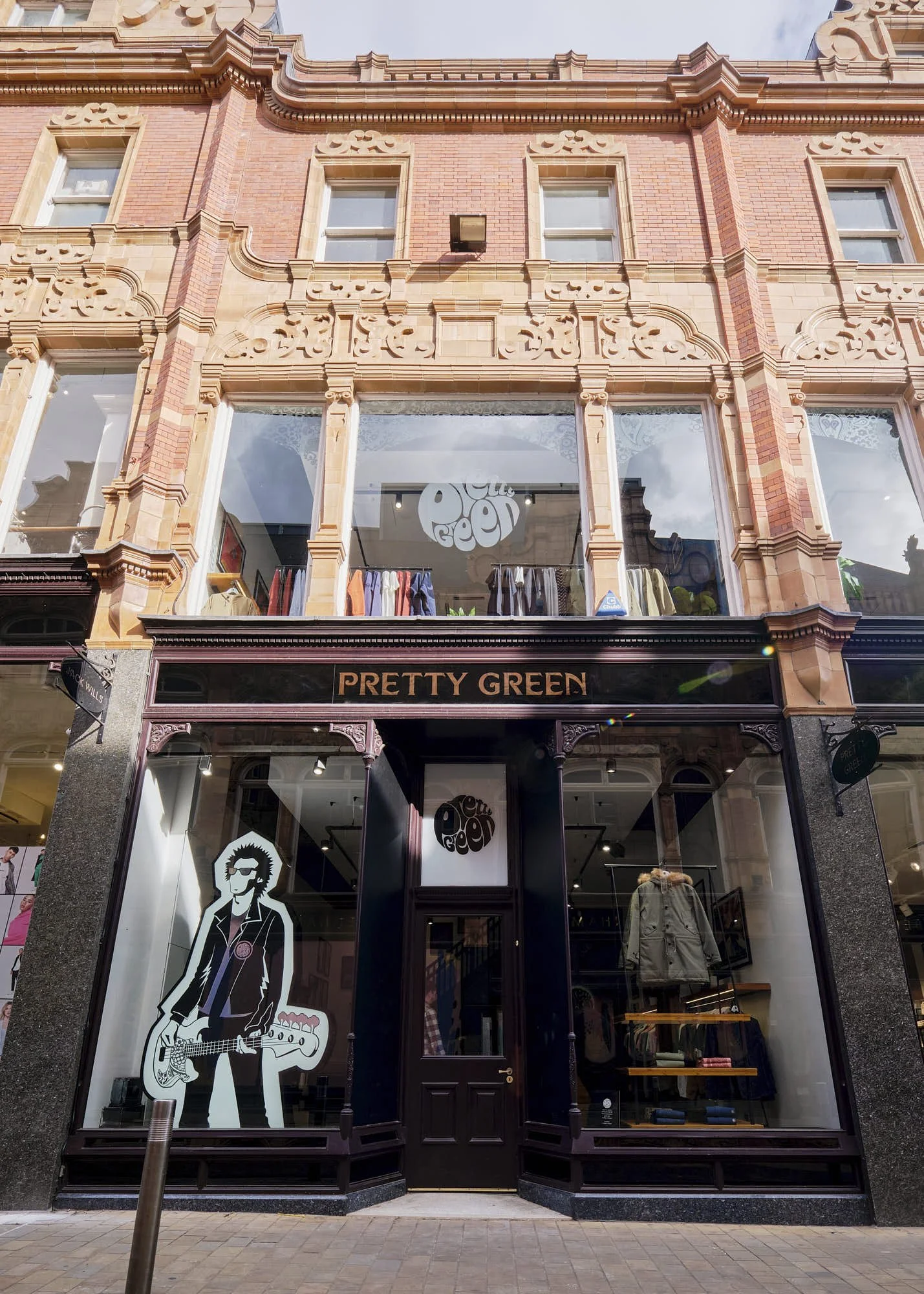 The storefront of Pretty Green clothing store with a glass window and door, displaying a black and white cartoon image of a person with glasses and an afro, holding a guitar. The building facade is made of ornate red and beige brickwork.