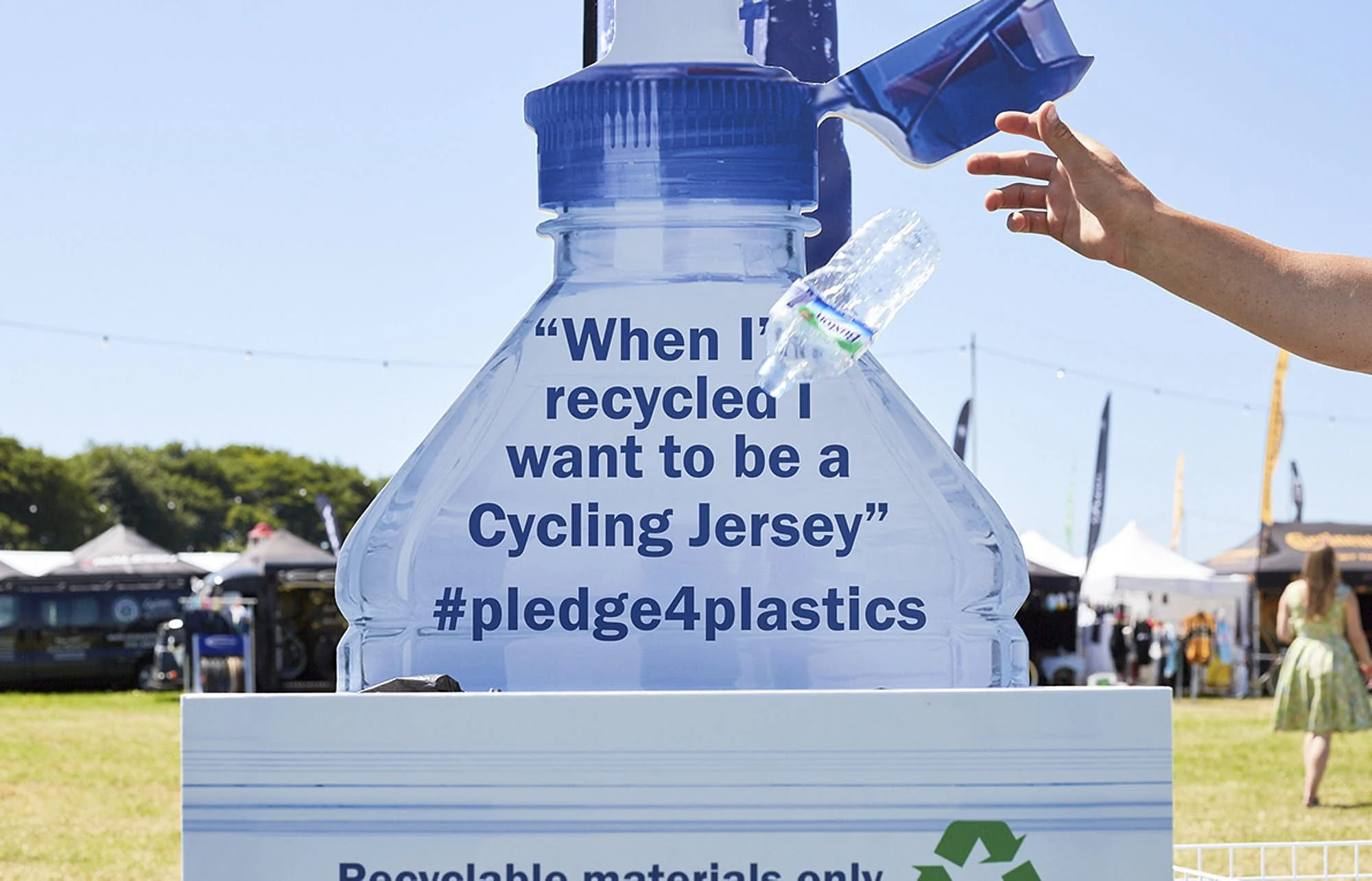 A large clear plastic water bottle with a blue cap and a hand placing a plastic water bottle into it. The bottle has text that reads: "When I recycle I want to be a Cycling Jersey" and includes hashtags #pledge4plastics and #recycling.