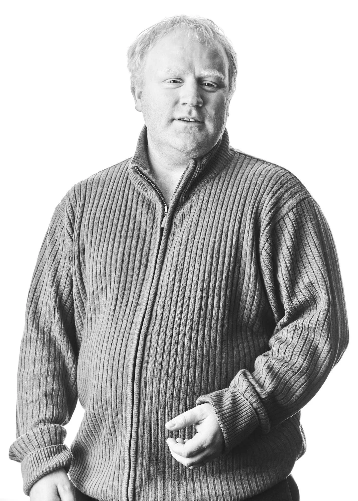 Black and white photo of a man with curly hair wearing a zip-up ribbed sweater, standing against a plain background.