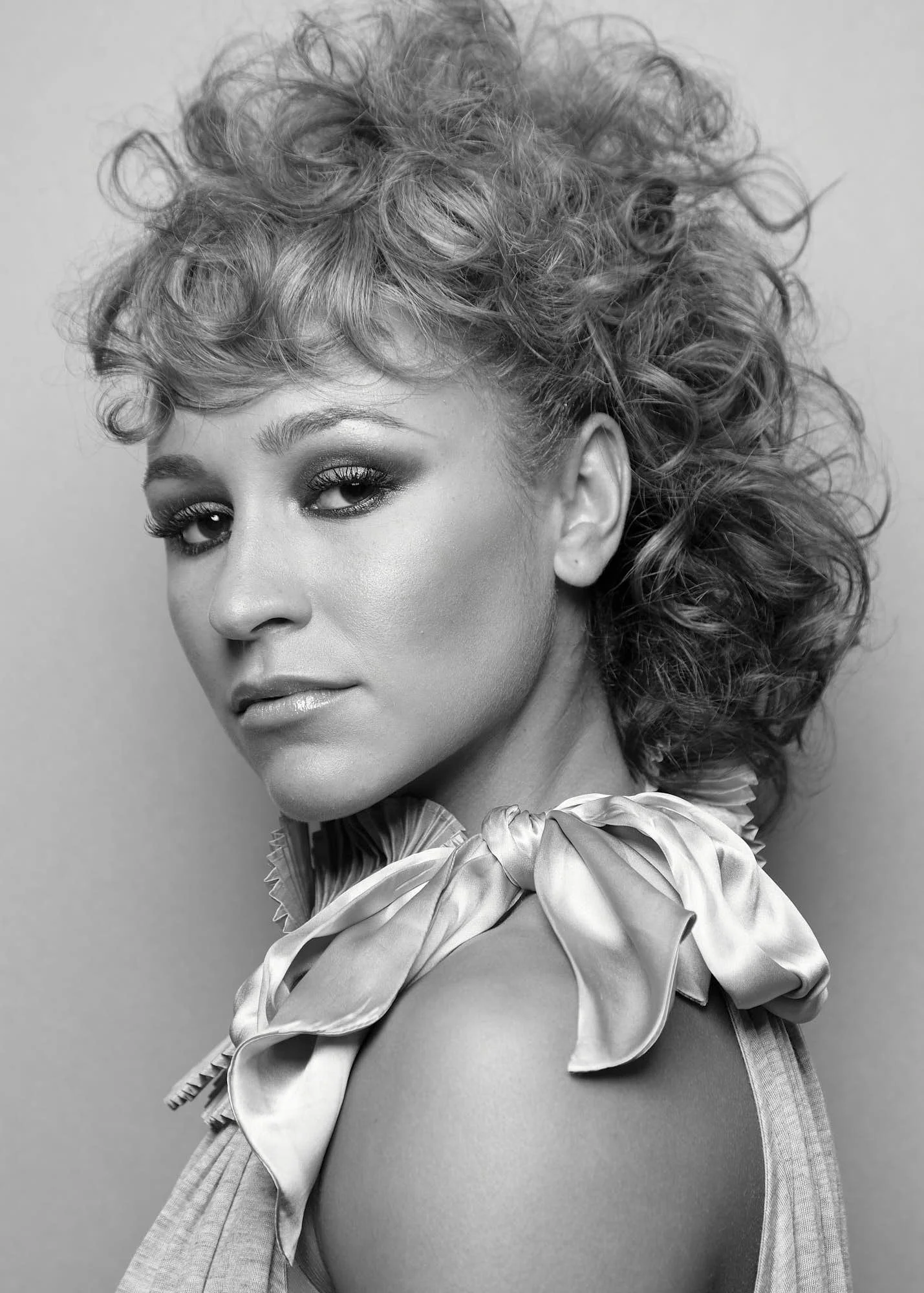Black and white photo of a woman with curly hair, makeup, and wearing a ruffled blouse with a bow around her neck.