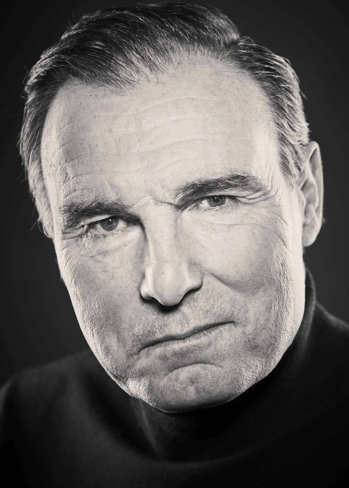 Close-up of a middle-aged man's face in black and white, featuring slicked-back hair, intense eyes, and a serious expression.