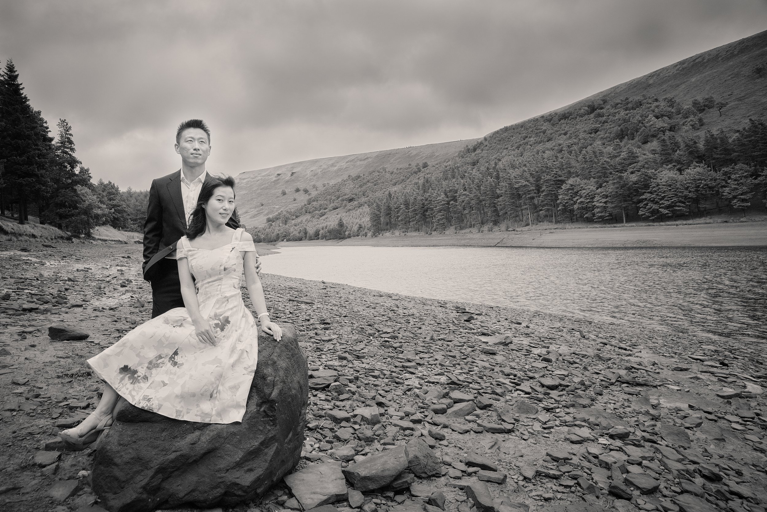 A black-and-white photo of a man and woman outdoors by a lake with hills and trees in the background. The woman is sitting on a large rock wearing a dress, and the man stands behind her in a suit, both looking at the camera.