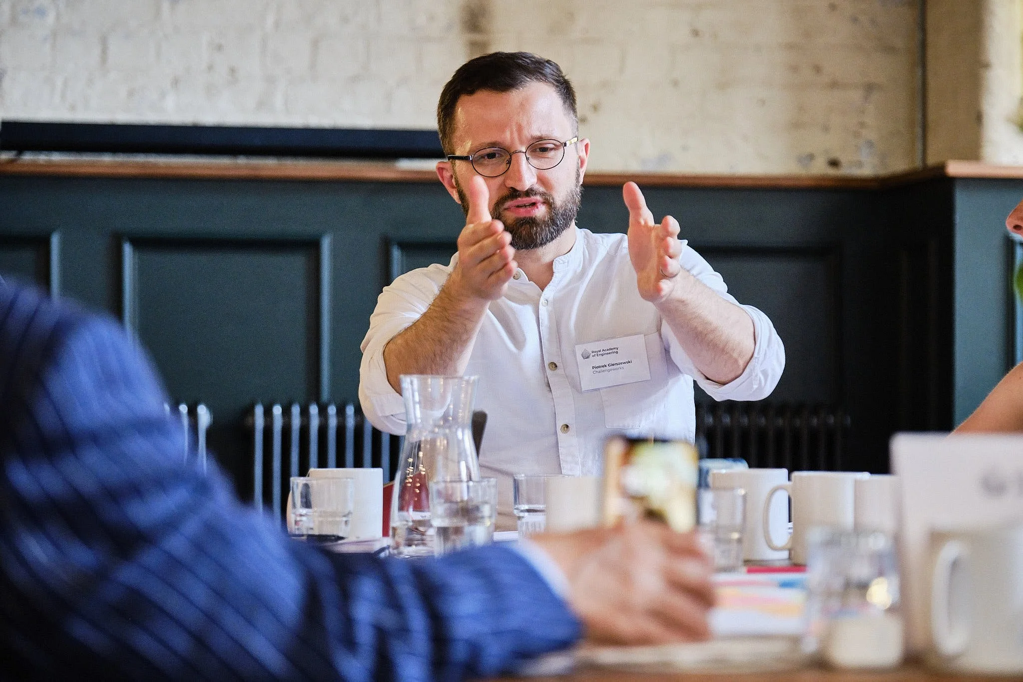 Man with glasses and beard gesturing during conversation at a table, in a professional or casual setting, with glasses, water pitcher, and decorative items on the table.