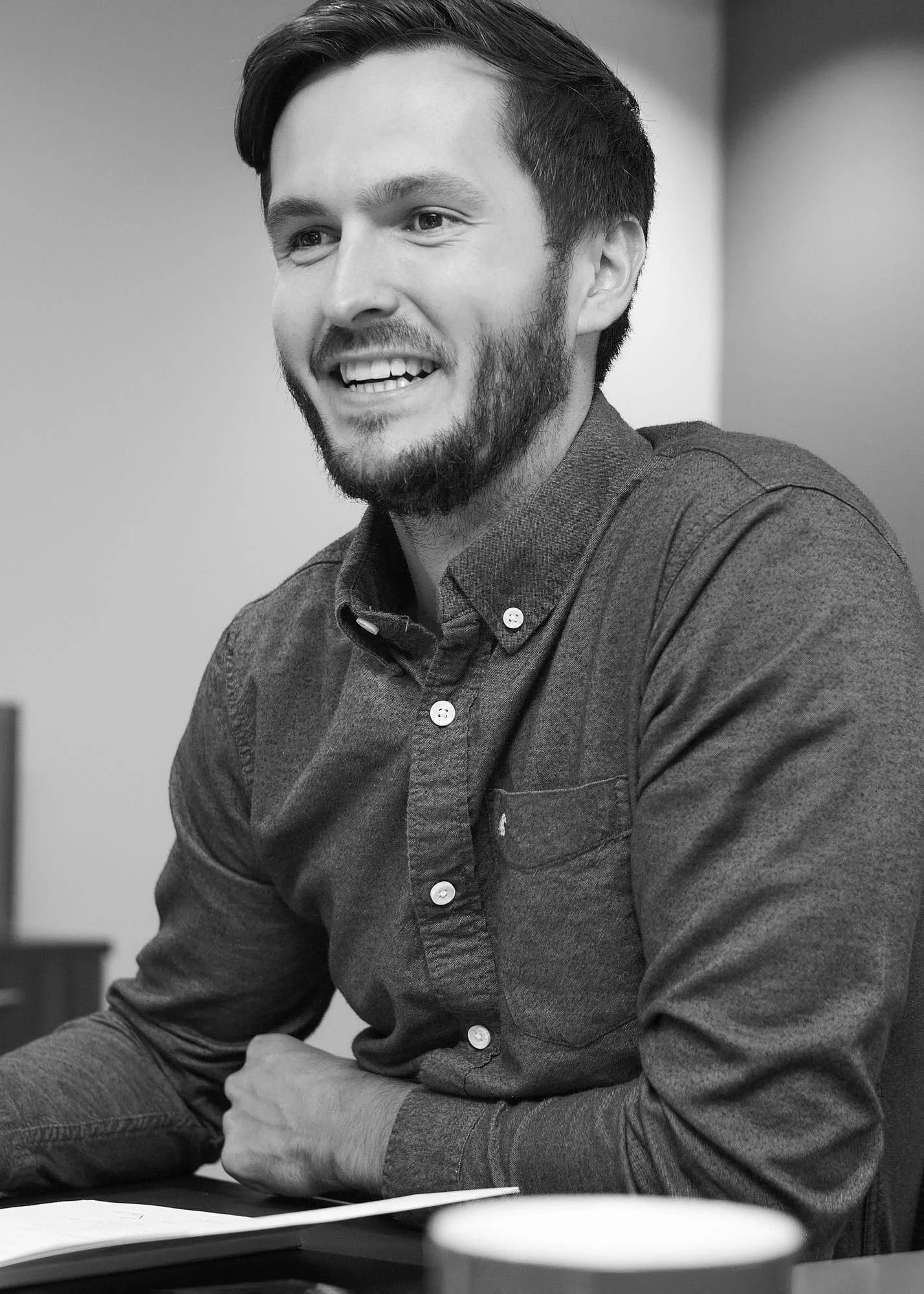Black and white photo of a young man with dark hair and a beard, smiling, sitting at a table with a book and a coffee mug.
