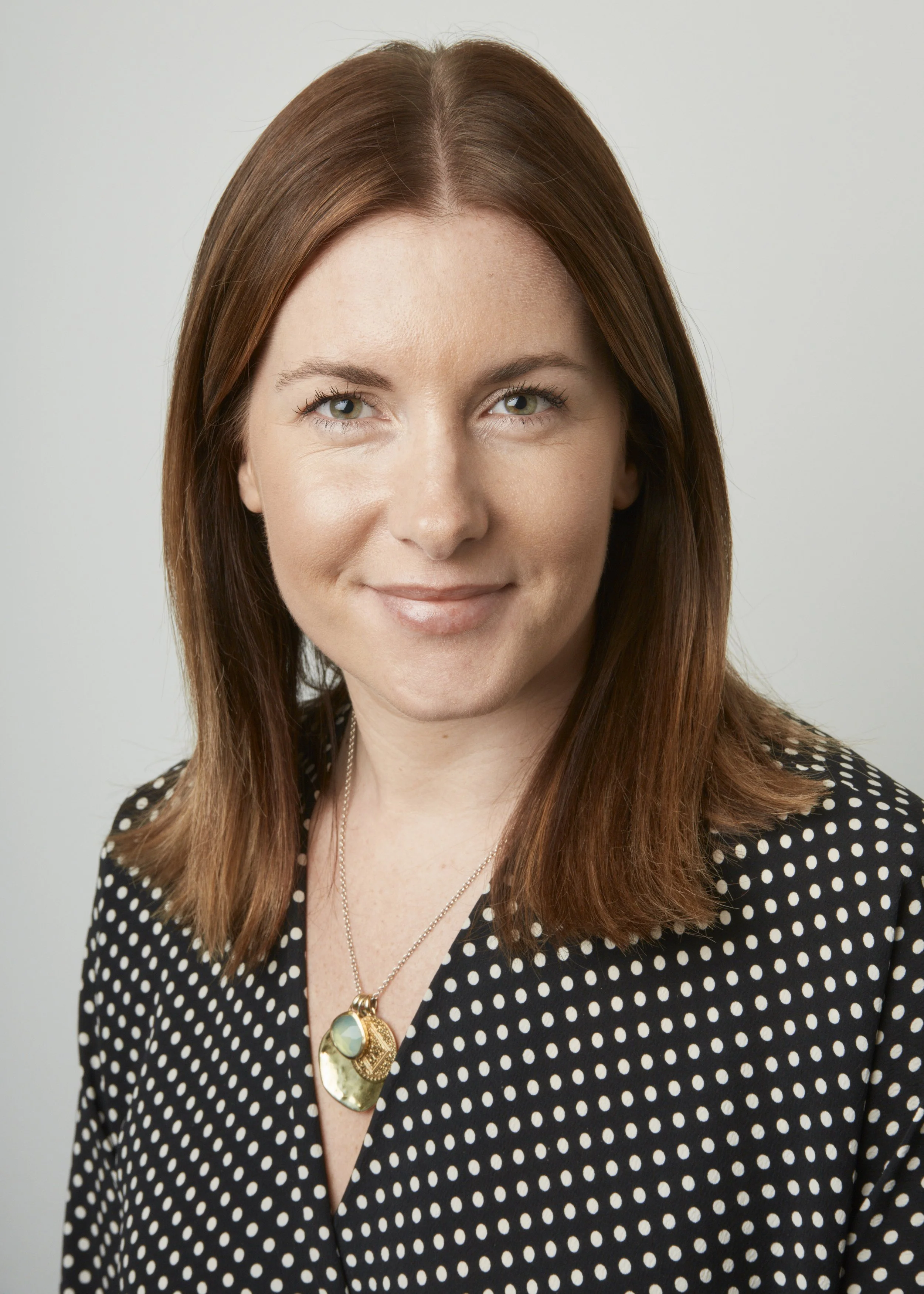 A woman with brown hair and blue eyes wearing a black dress with white polka dots, a gold necklace with a large oval pendant, and a gold chain necklace. She is smiling slightly and looking at the camera against a light, neutral background.
