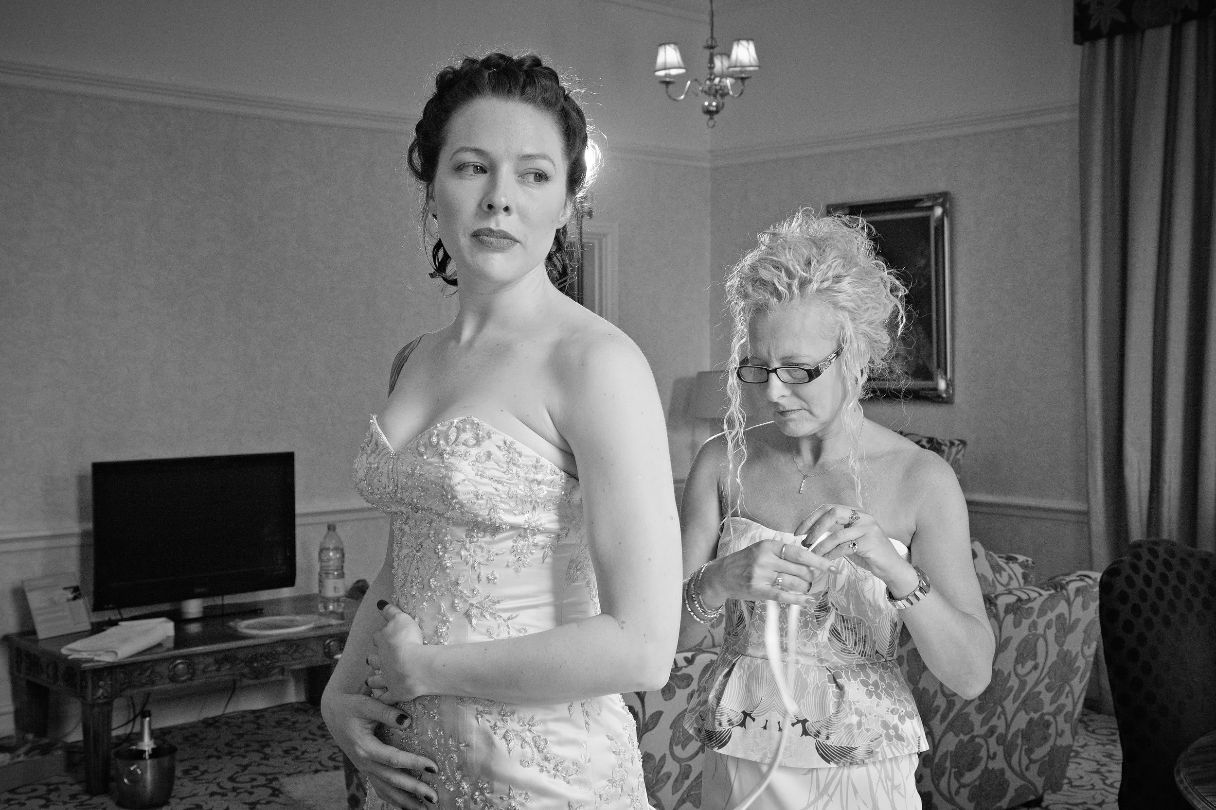 Black and white photo of a woman in a wedding dress standing and looking to the side while another woman behind her adjusts her dress in a room decorated with curtains, framed pictures, and a chandelier.